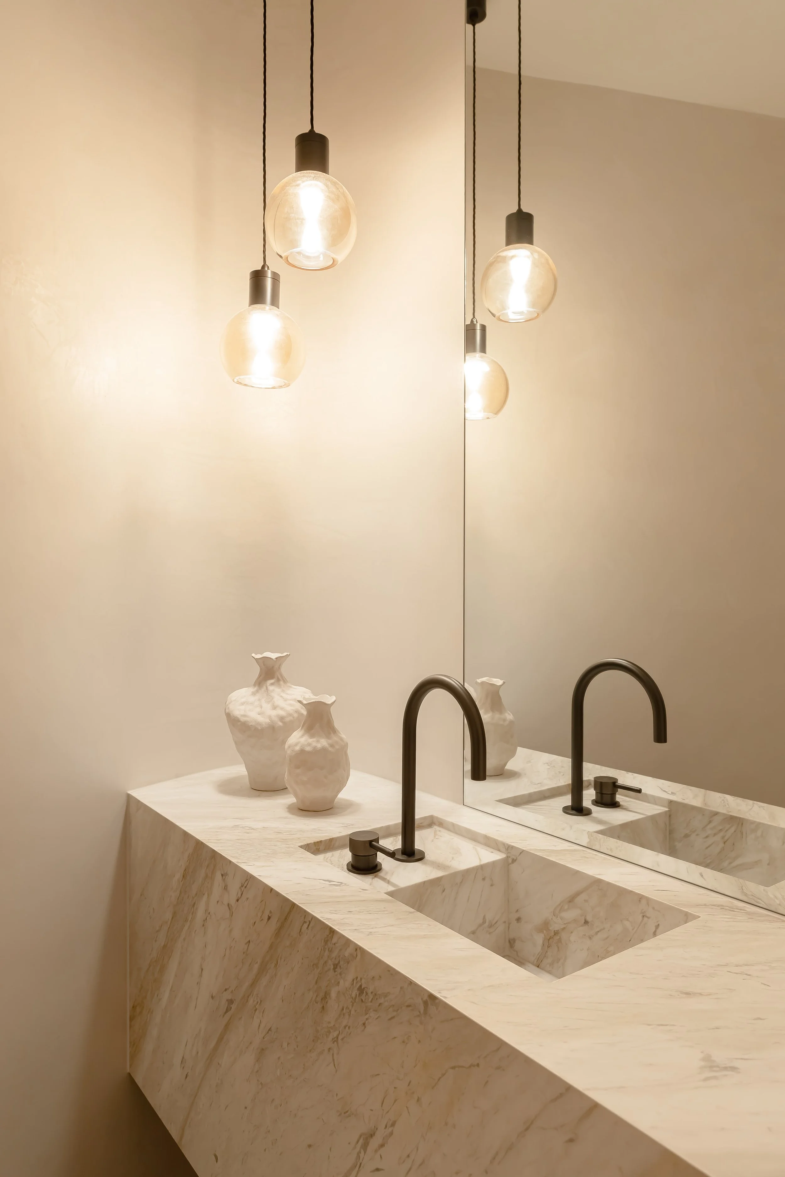 A bathroom vanity with a marble countertop, two black faucets, white vases, and hanging pendant lights reflected in a large mirror.