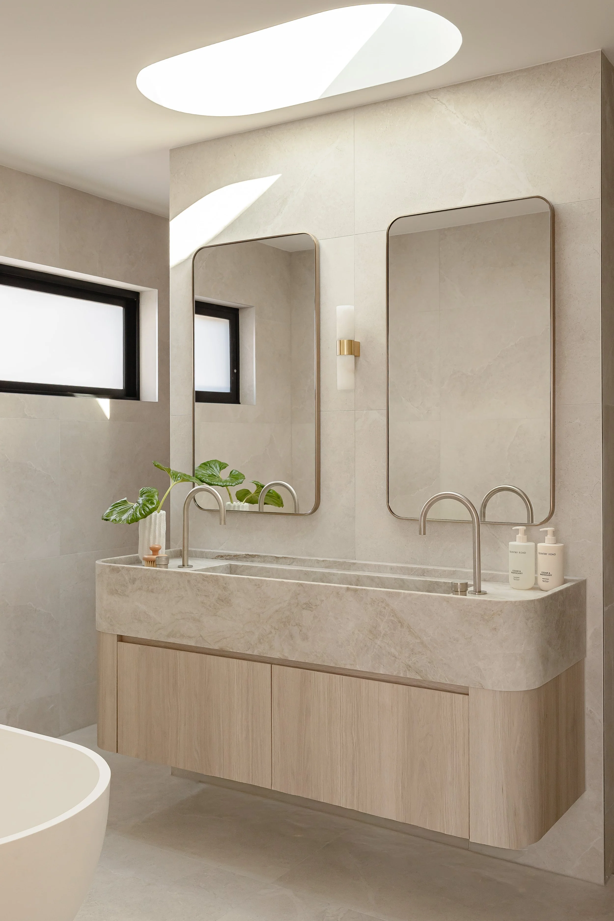 Modern bathroom vanity with double sinks, two mirrors, a potted plant, natural light from rectangular windows, and minimalistic decor.