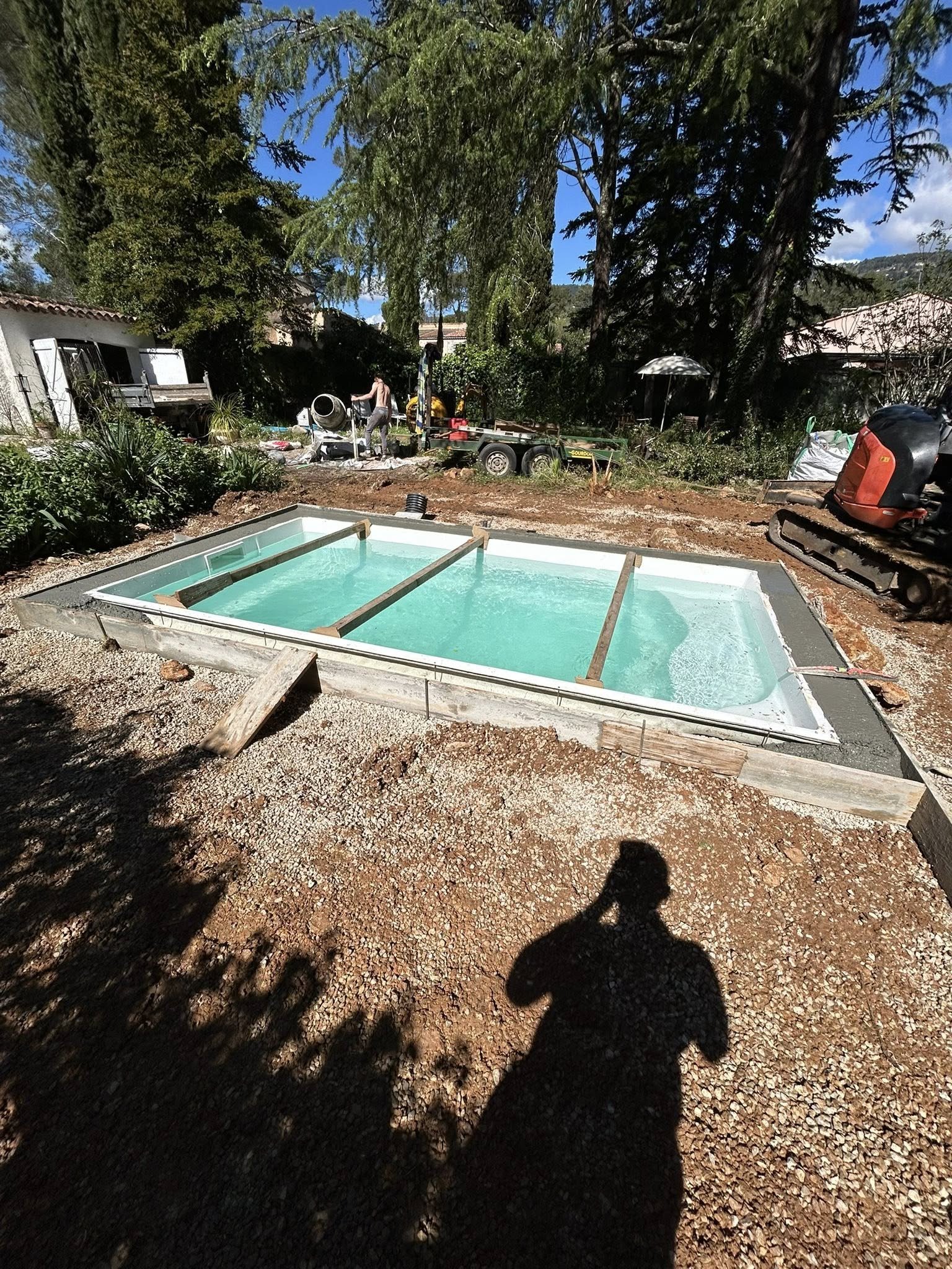 Une piscine en construction dans un jardin, avec du bavoir d'eau et des outils, sous un ciel bleu avec des arbres en arrière-plan.