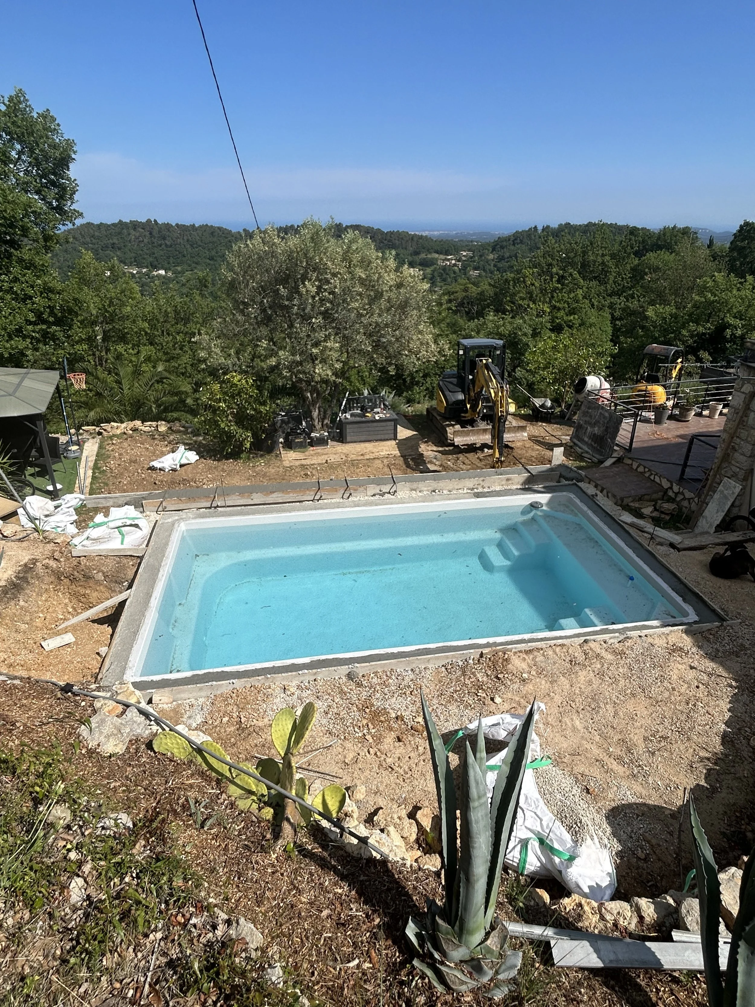 Une petite piscine vide en cours d'installation dans un jardin. En arrière-plan, des travaux avec des mini-pelles, des outils et du matériel de construction. Le paysage est verdoyant avec des arbres et une vue sur une région montagneuse et l'océan au loin.
