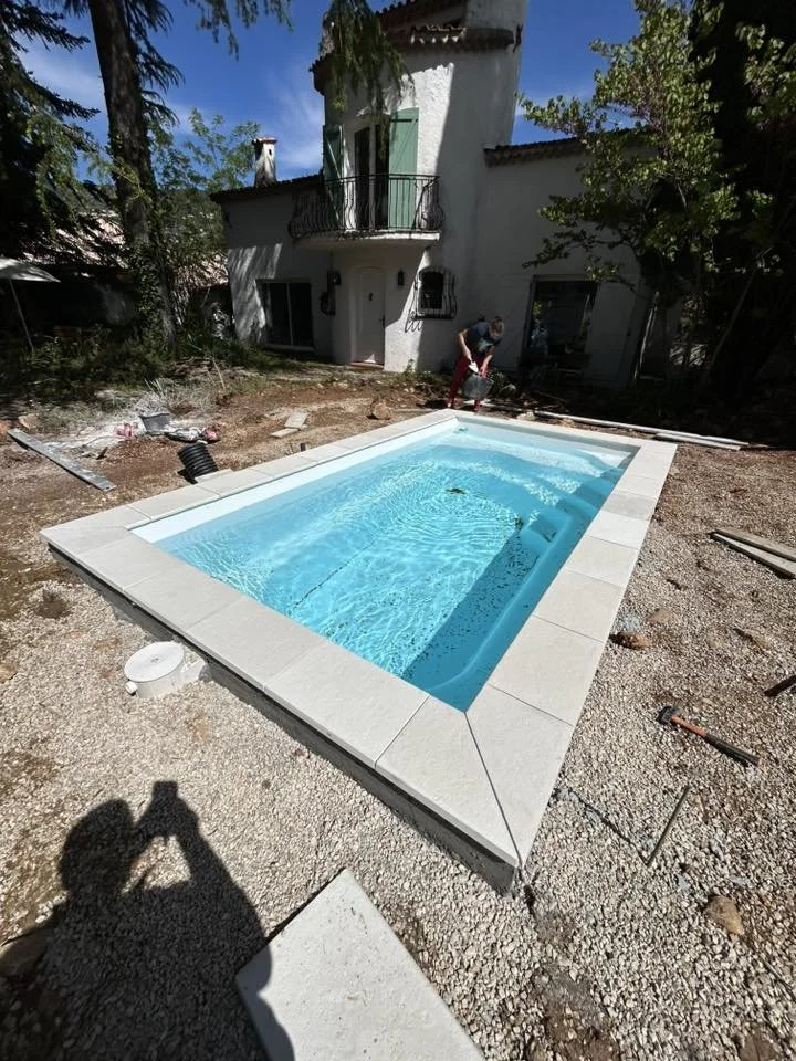 Une piscine en construction devant une maison blanche avec un balcon en fer forgé, entourée de jardin et de travaux en cours.