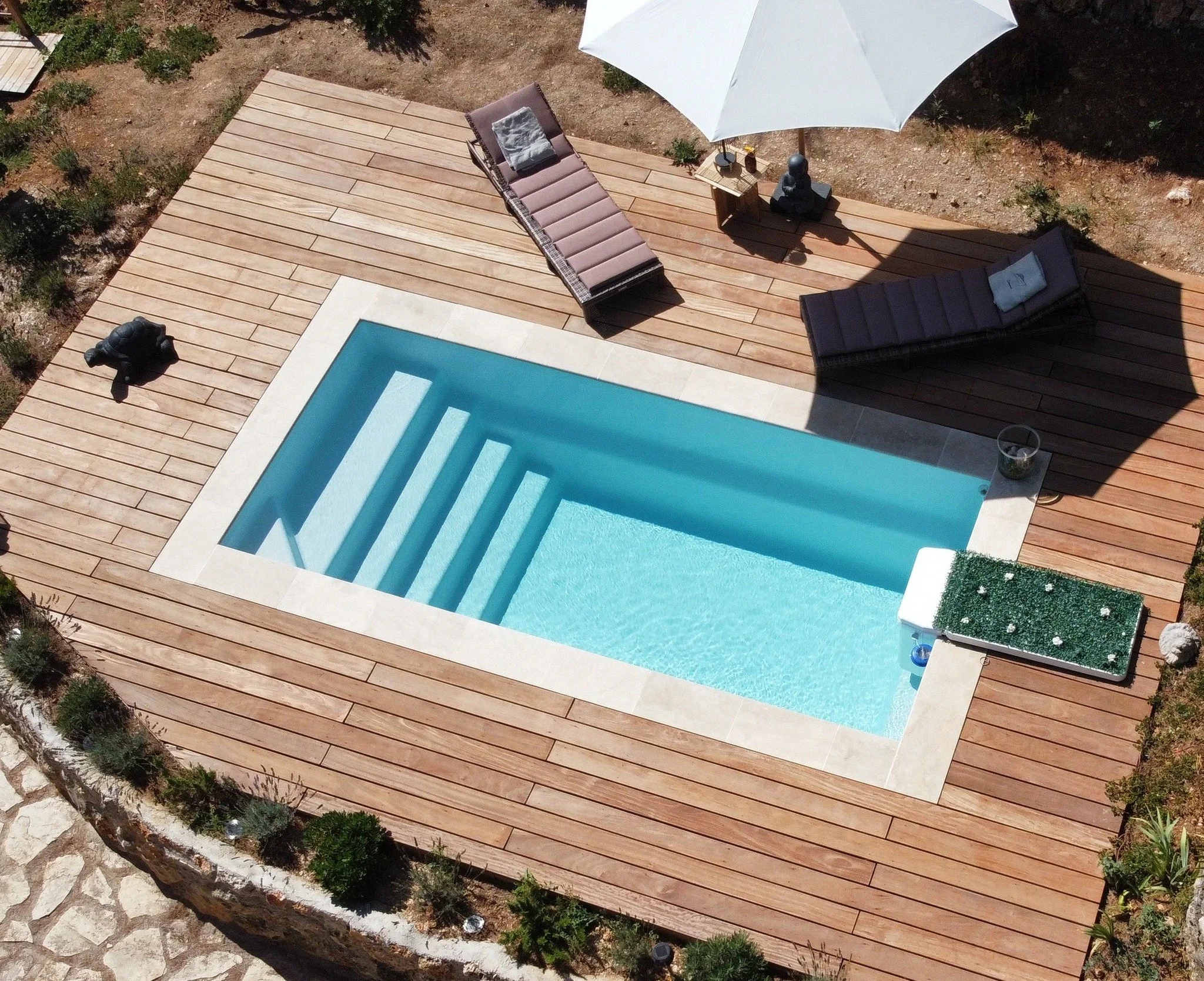 Vue aérienne d'une piscine rectangulaire avec des marches en pierre blanche, entourée d'une terrasse en bois, avec deux transats, un parasol blanc, et des objets de décoration.