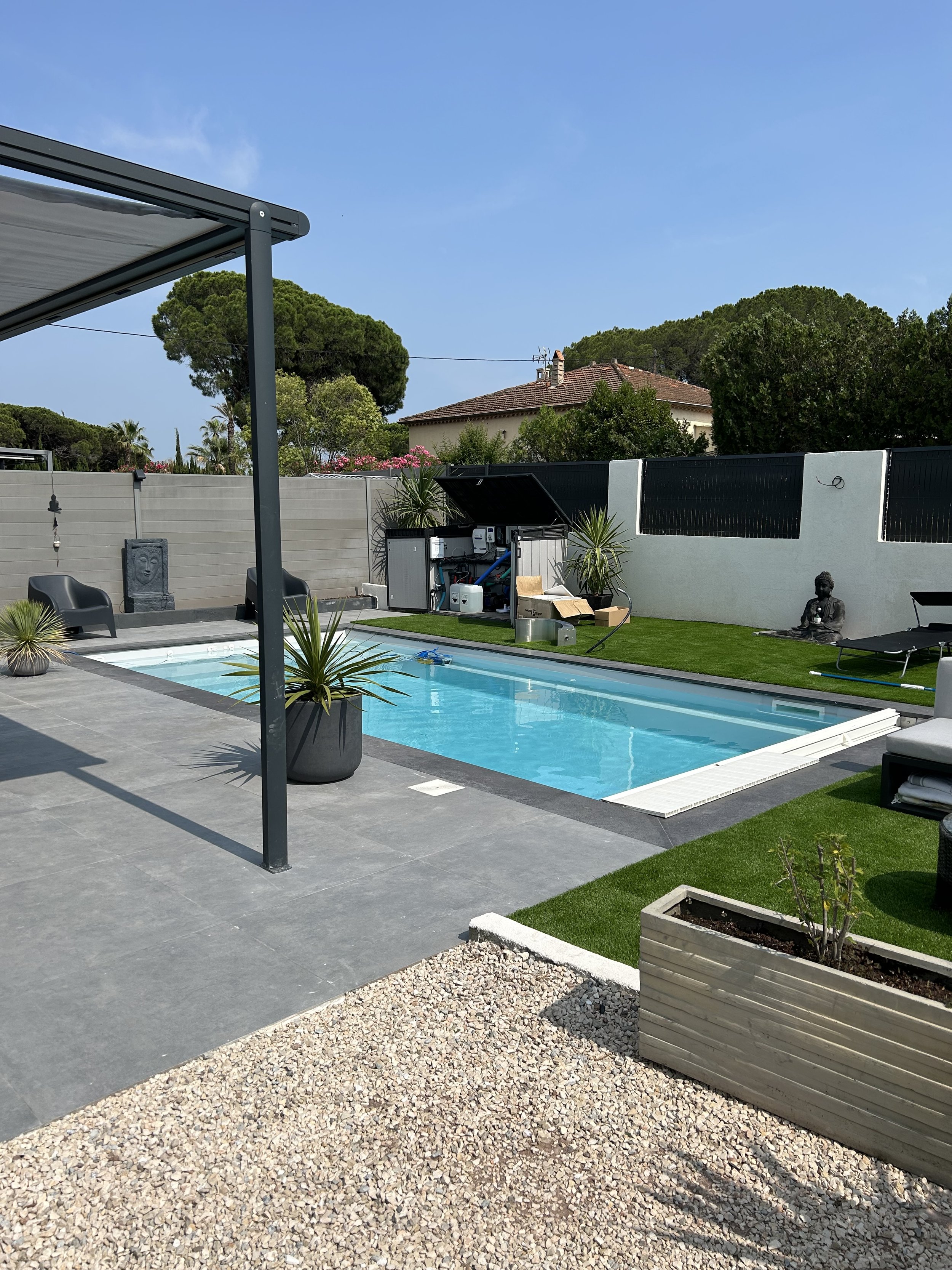 Piscine extérieure avec mobilier de jardin, plantes en pot, statue de Bouddha, jardin bien entretenu, et maison en arrière-plan sous un ciel bleu.