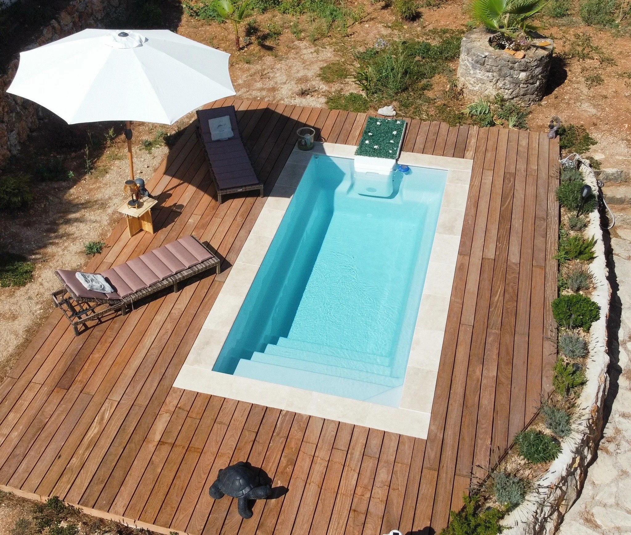 Piscine rectangulaire avec escalier, entourée d'une terrasse en bois, avec chaises longues, parasol blanc, et plantes décoratives.