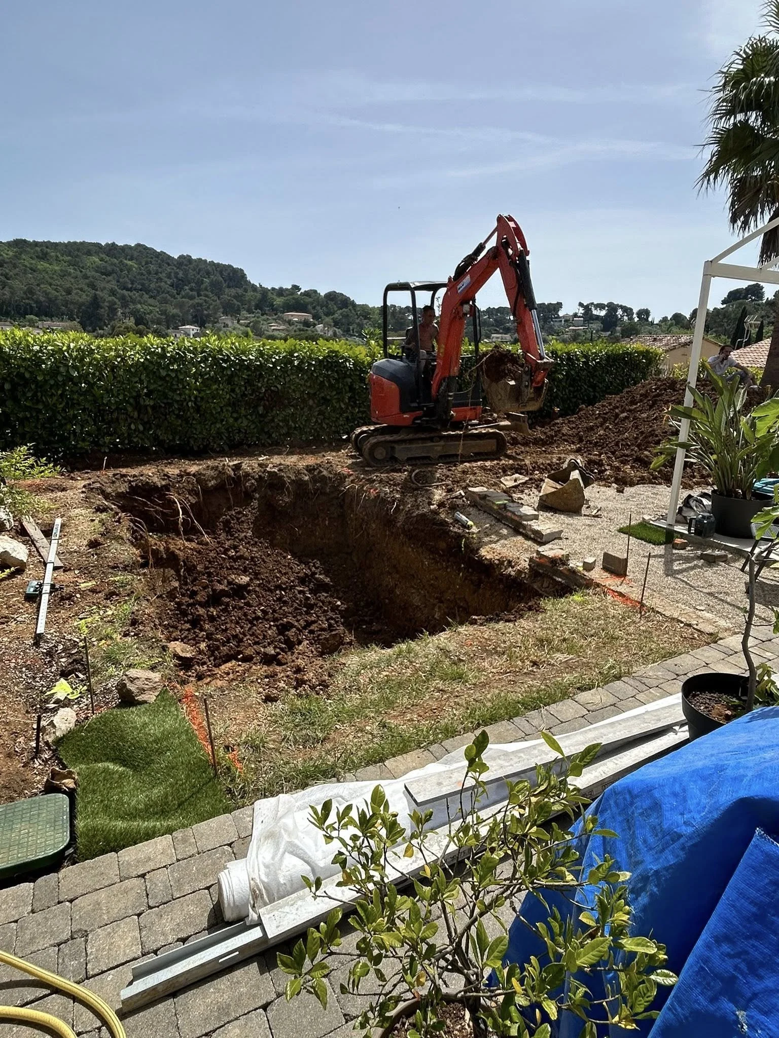 Une excavation en cours dans un jardin, avec une personne utilisant une petite excavatrice orange. Le ciel est dégagé et il y a des collines verdoyantes en arrière-plan.