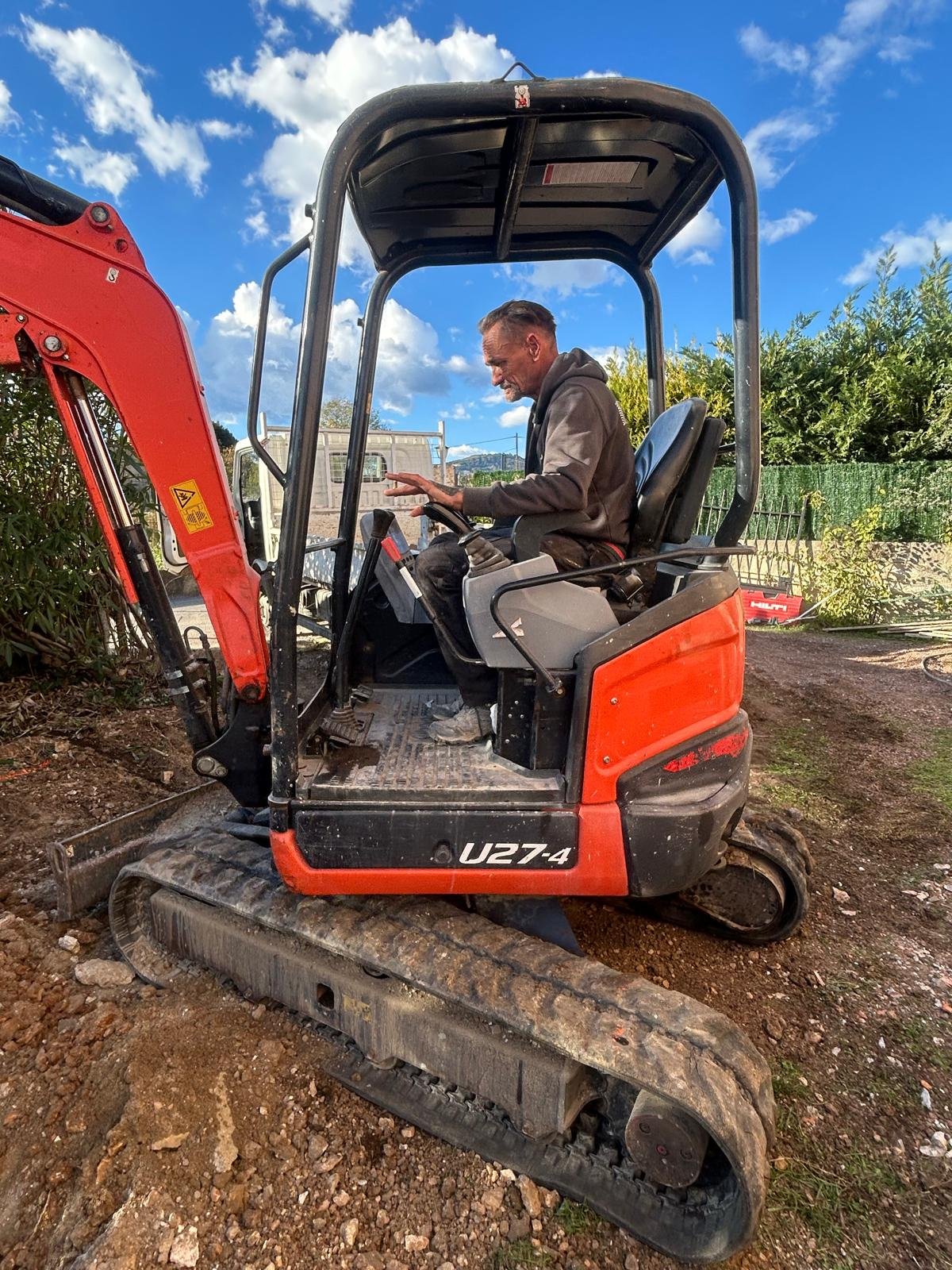 Un homme assis dans une petite excavatrice de couleur orange et noire, travaillant à l'extérieur sur un terrain en terre avec des buissons en arrière-plan et un ciel bleu avec quelques nuages.