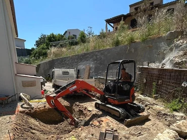 Maçonnerie avec une petite excavatrice rouge en action, creusant un trou sur un chantier à proximité de maisons, avec des bâtiments en arrière-plan et un ciel bleu clair.