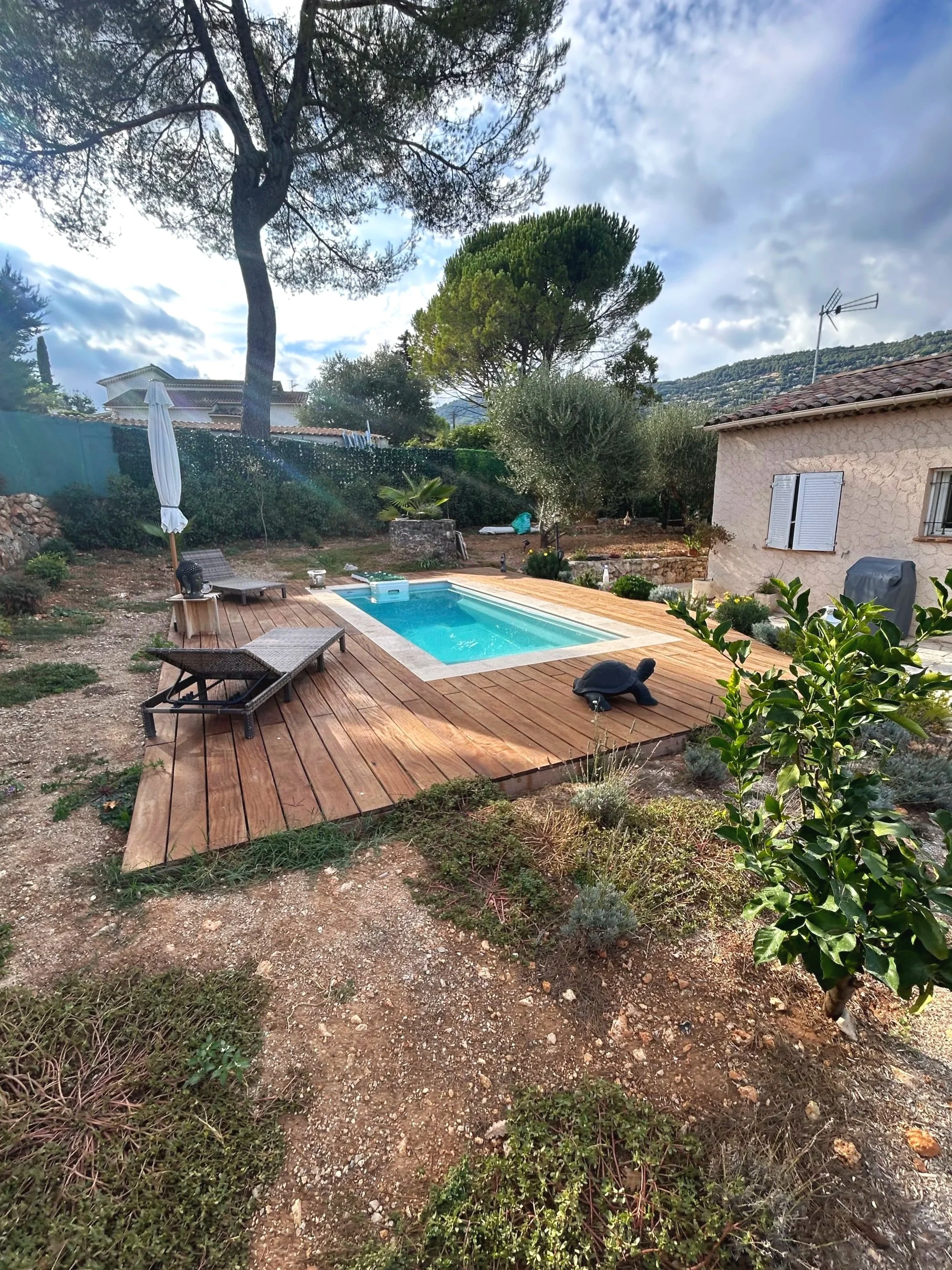 Une piscine dans un jardin entourée de plantes, avec un sol en bois autour, une chaise longue et un parasol fermé, une maison en pierre avec volets blancs, et un ciel nuageux avec quelques rayons de soleil.