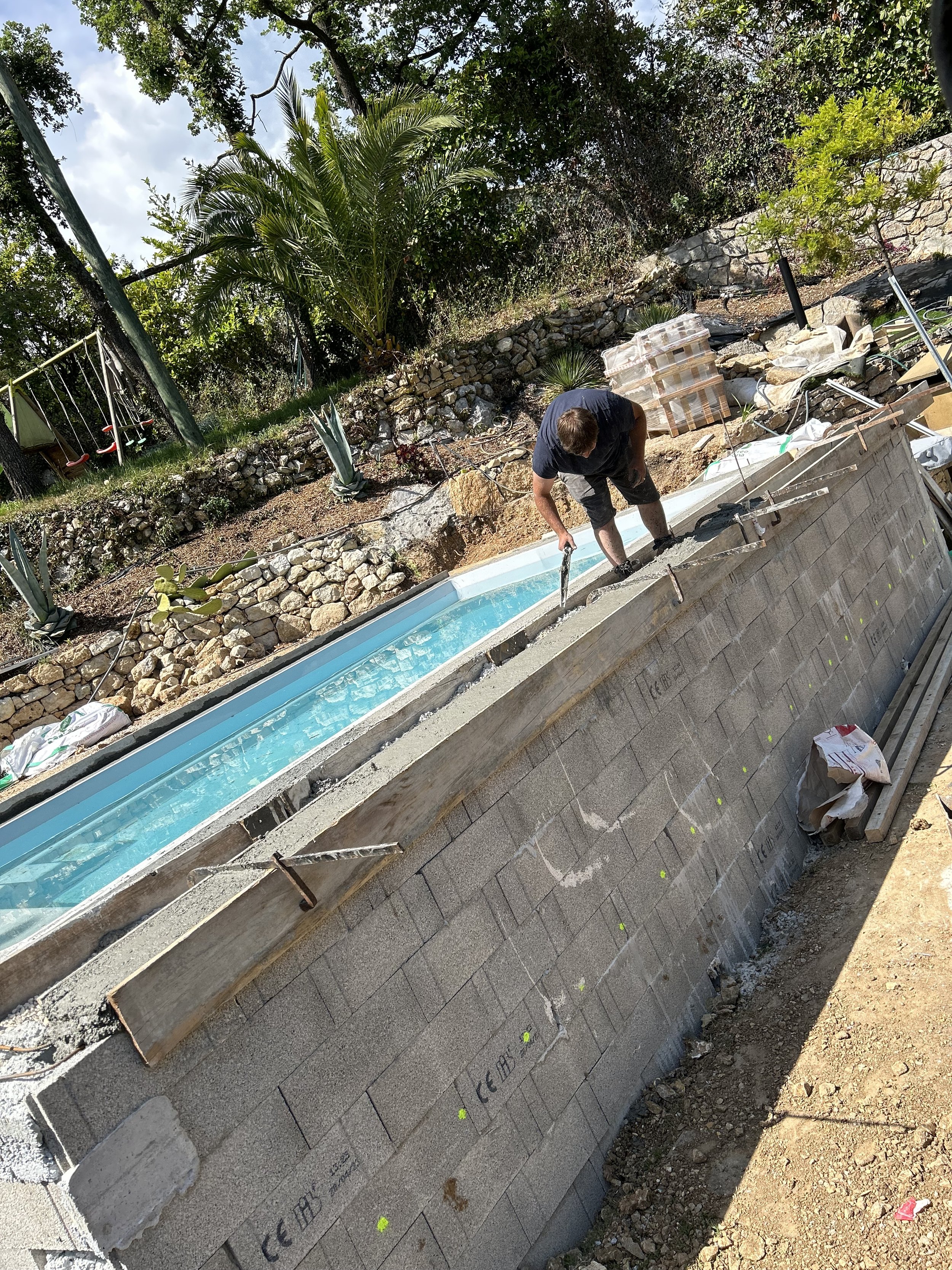 Un homme construit une piscine hors sol dans un jardin, entouré de rochers et de plantes tropicales, en utilisant des blocs de béton.