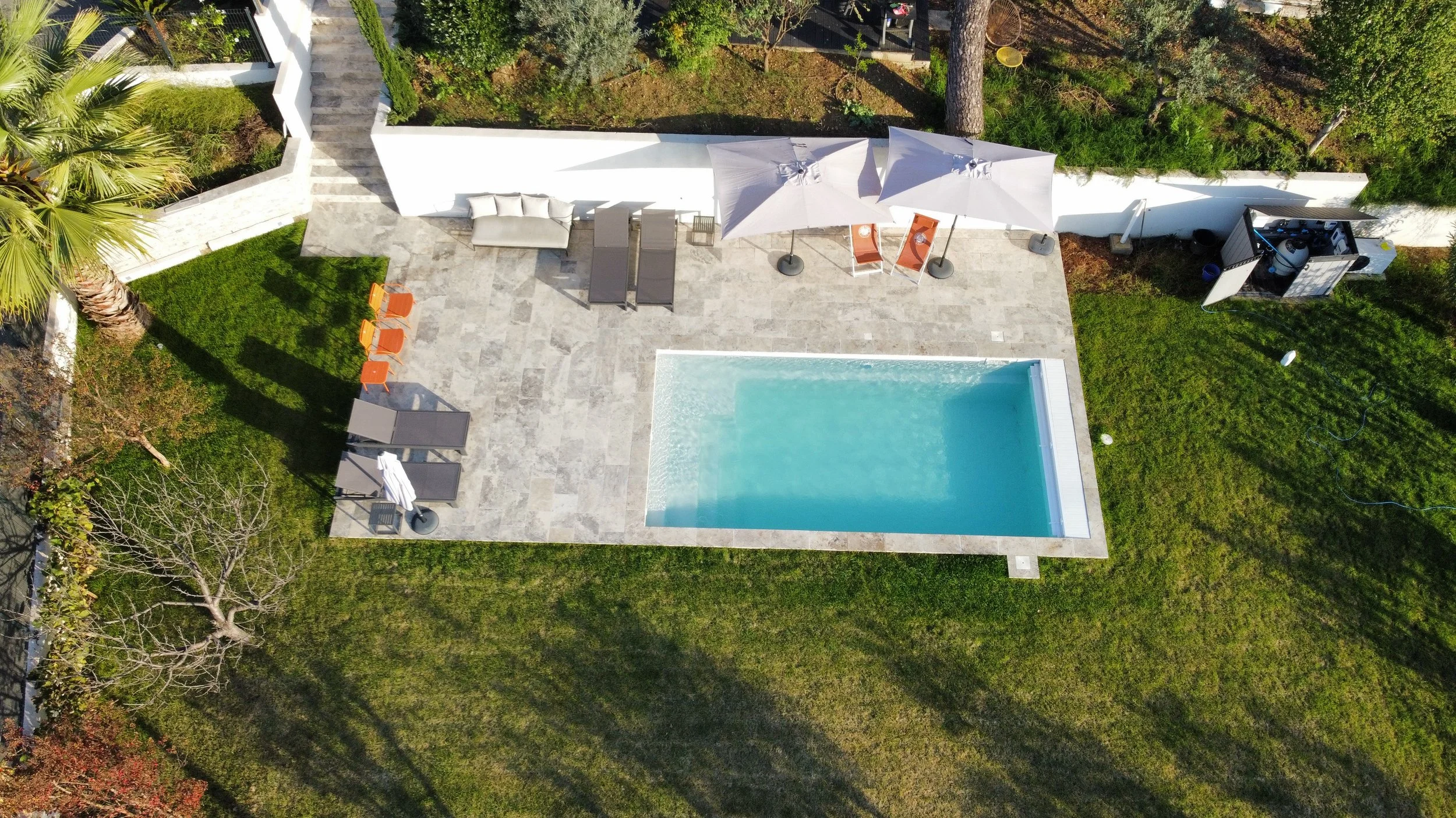 Une piscine dans un jardin avec des transats, parasols, et quelques meubles, vue d'une perspective aérienne.