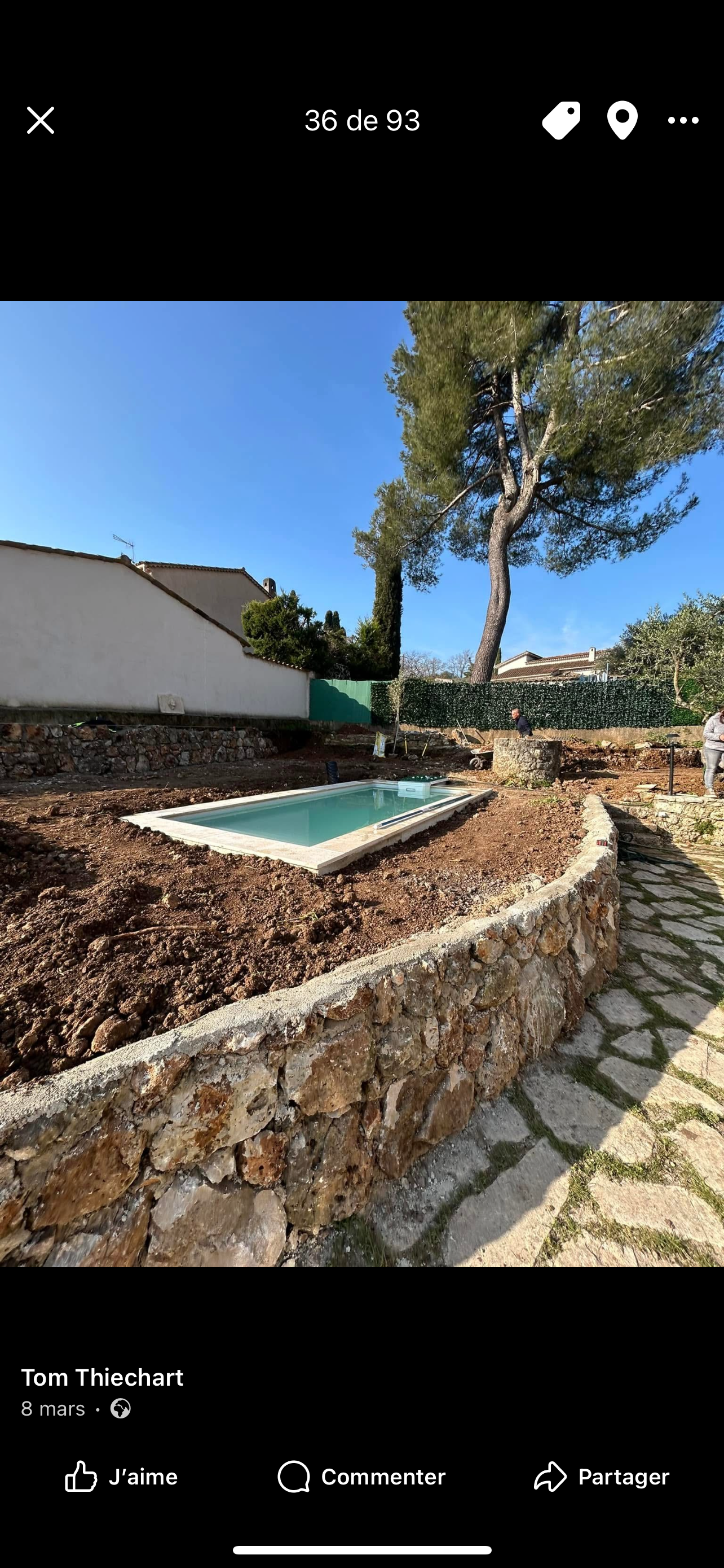 Piscine en construction dans un jardin avec une clôture en pierre et des arbres, ciel bleu.