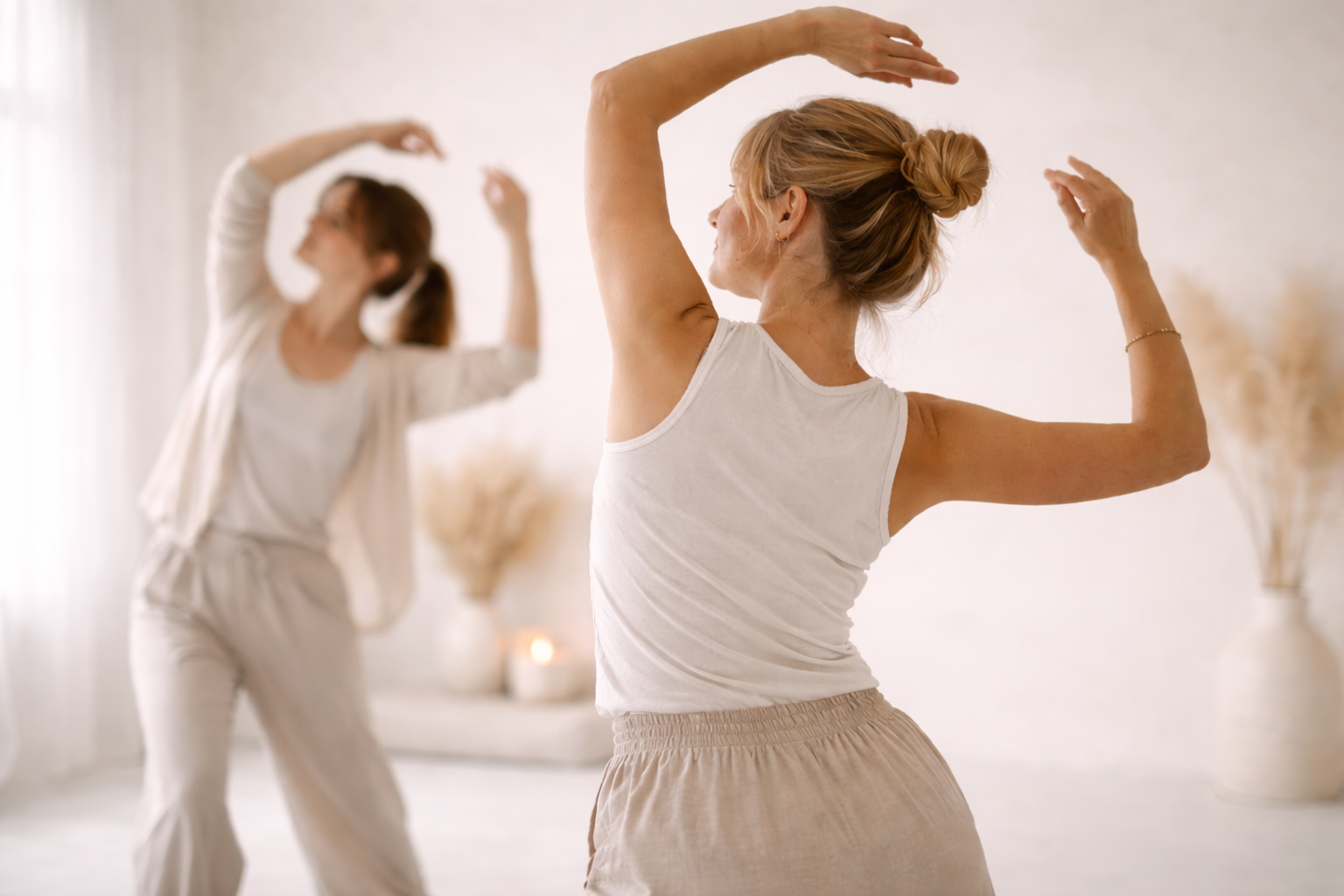 Two women practicing yoga or stretching in a bright, minimalistic room with beige decor and dried plants.