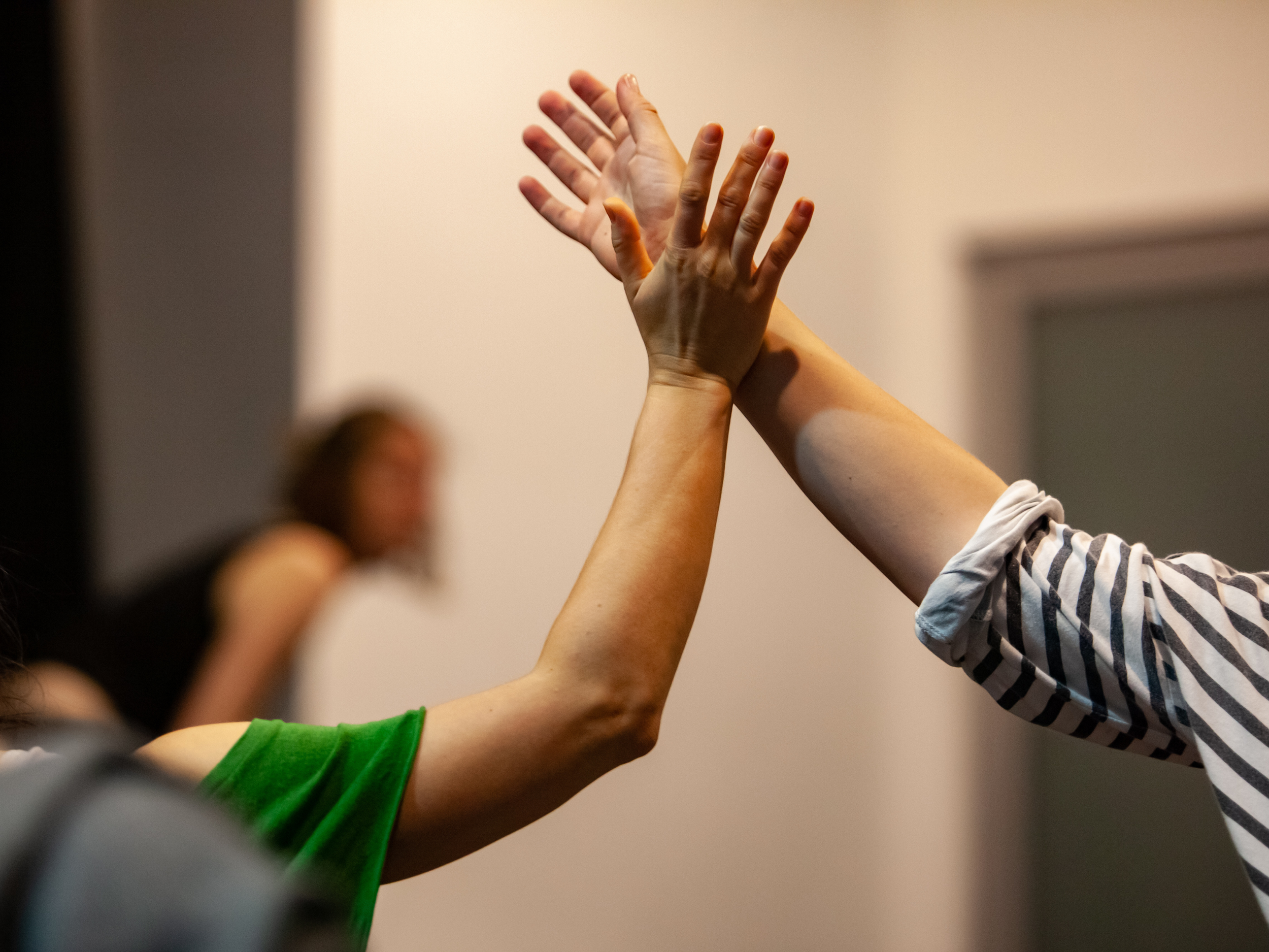 Two people high-fiving, with only the arms and hands visible, one wearing a green shirt and the other in a striped shirt, in a blurred indoor setting.