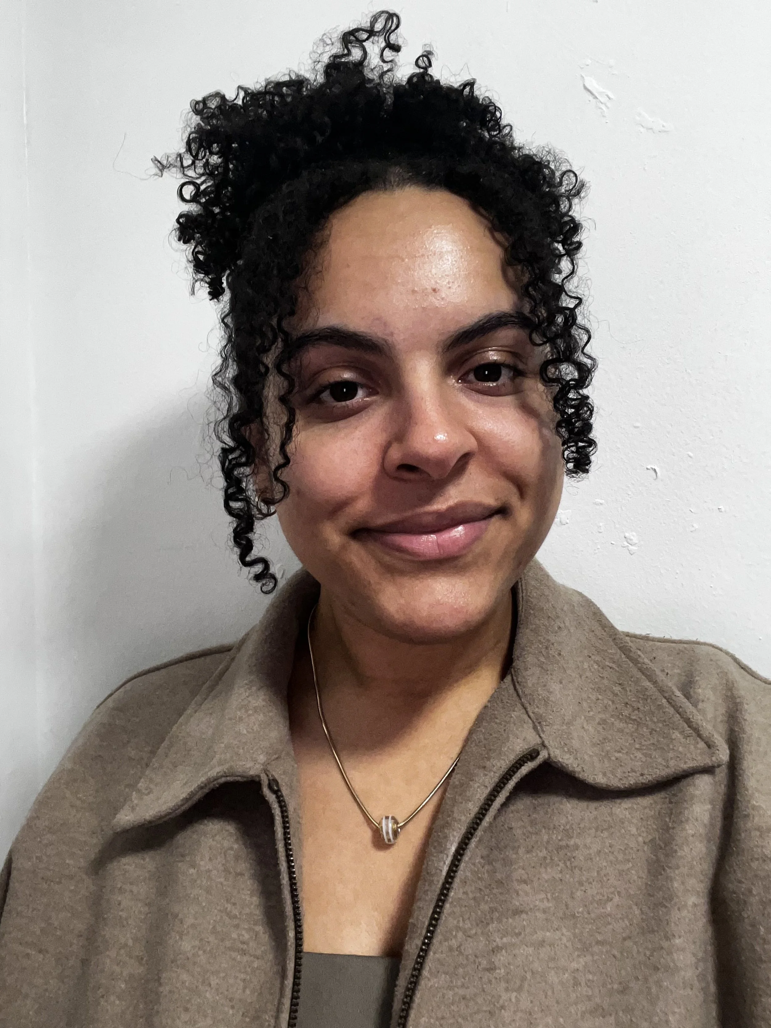 A woman with curly black hair smiling at the camera, wearing a brown jacket and a silver necklace, standing against a white wall.