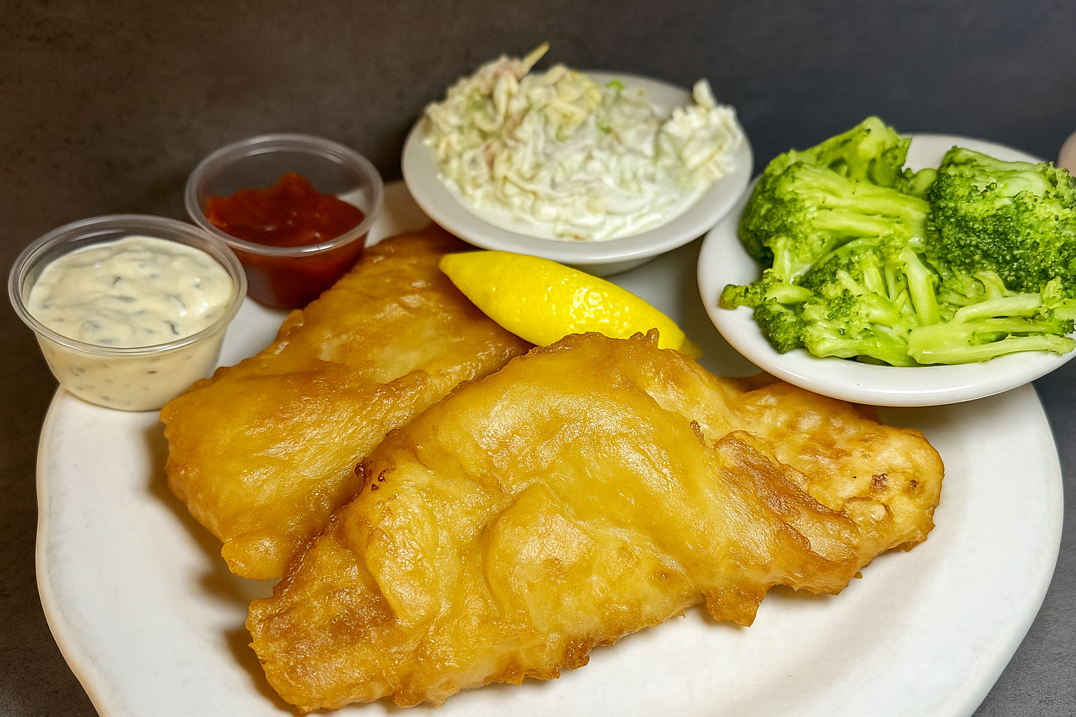 A plate of fish and chips with sides of broccoli, coleslaw, tartar sauce, ketchup, and lemon.