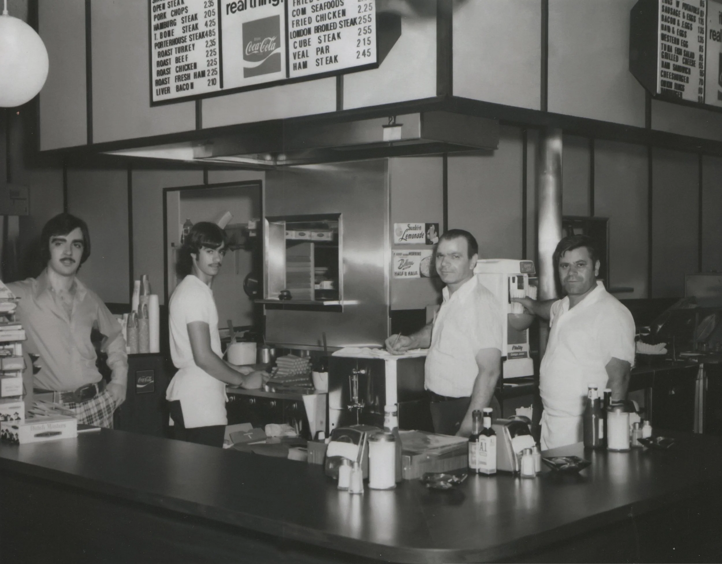 Black and white photo of a retro fast-food restaurant counter with two male employees in white uniforms, one male employee behind the counter, and two female employees, one with a plaid skirt, working behind the counter. Men are looking towards camer