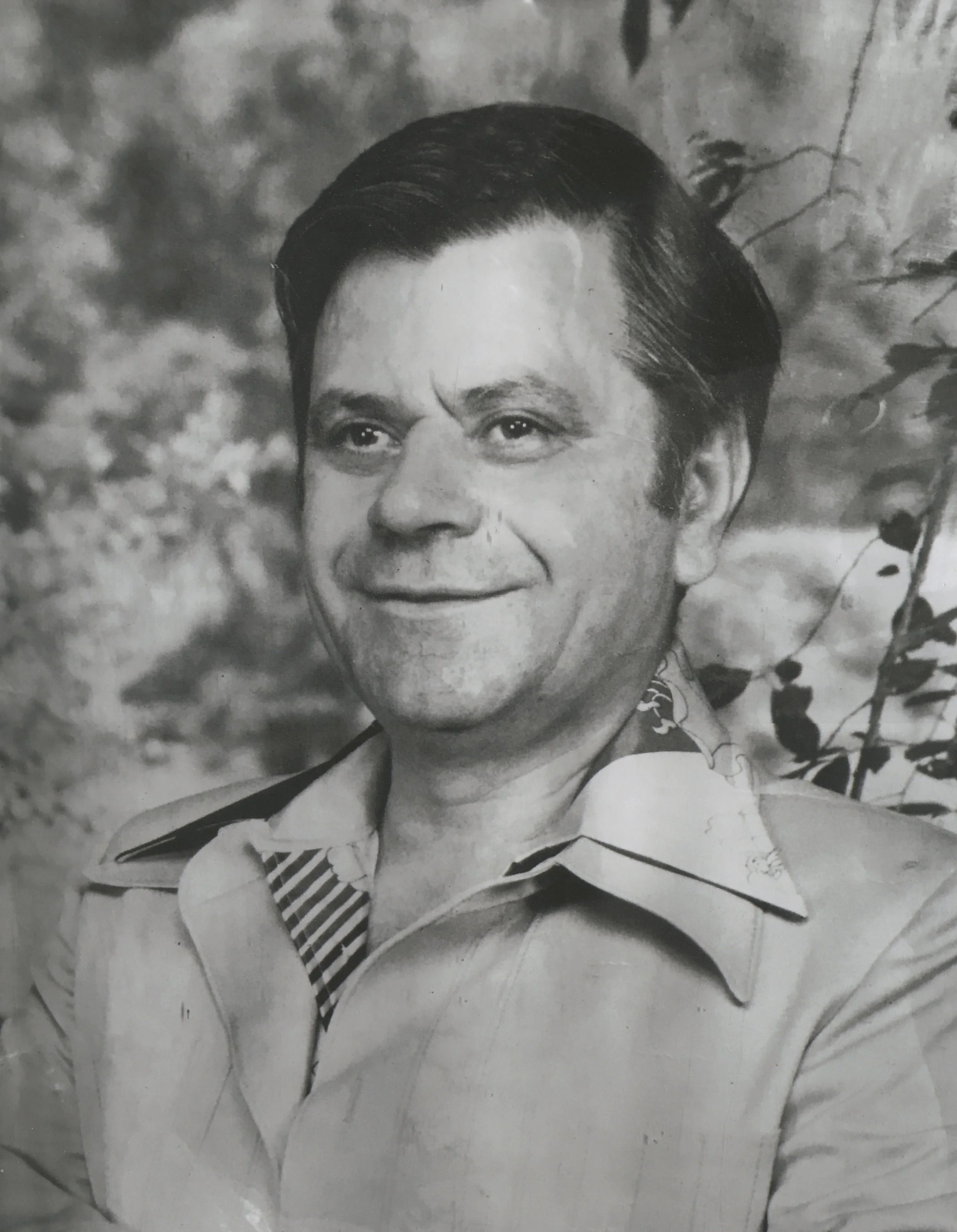 Black and white portrait of a smiling man with styled dark hair, wearing a collared shirt with a patterned inner collar, in front of a nature background.