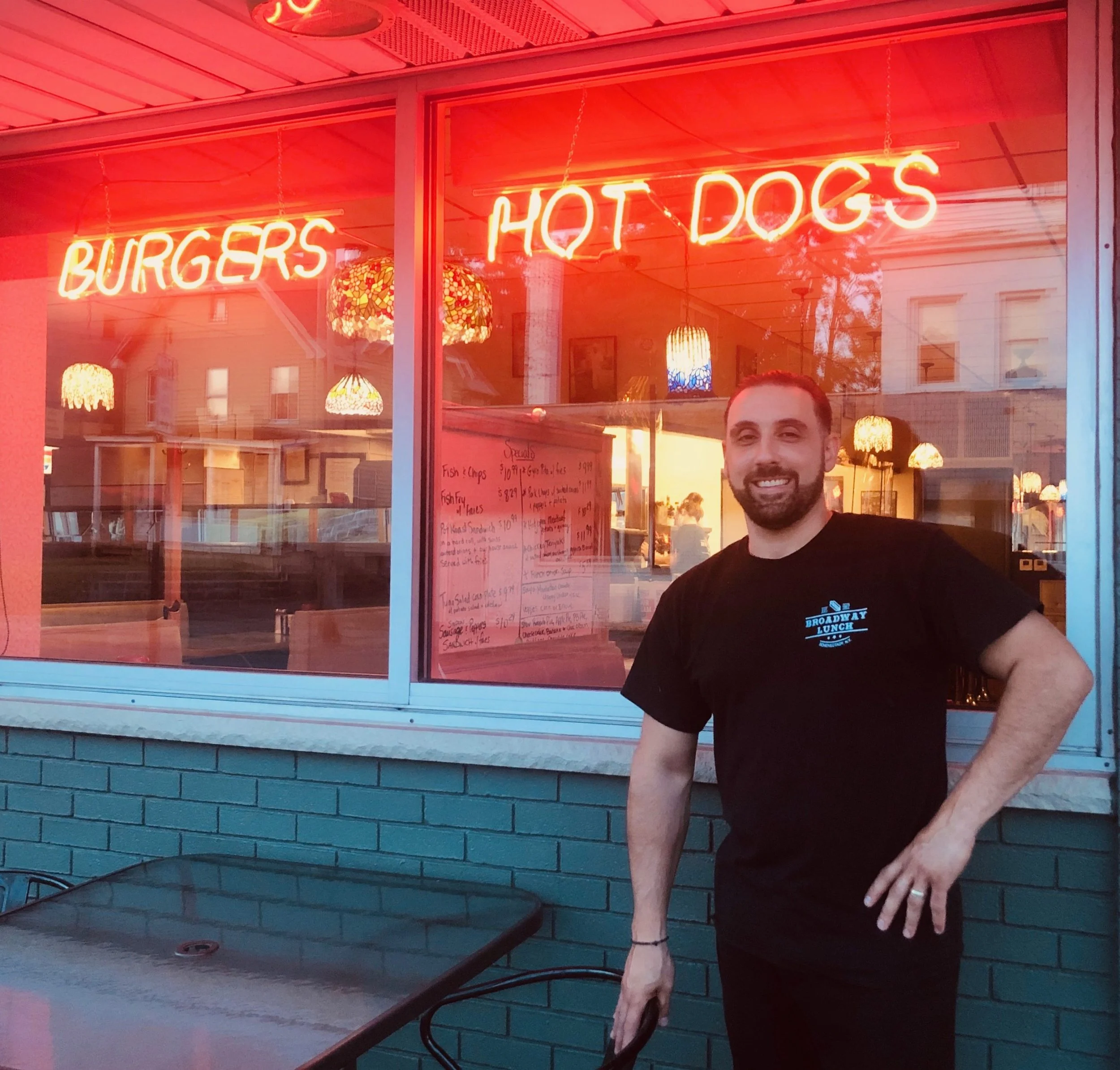 A man standing outside a restaurant with a neon sign displaying 'BURGERS' and 'HOT DOGS' in red lights, wearing a black T-shirt with a blue and white logo, smiling with one hand on his hip, in front of a window with interior lighting and hanging lamp