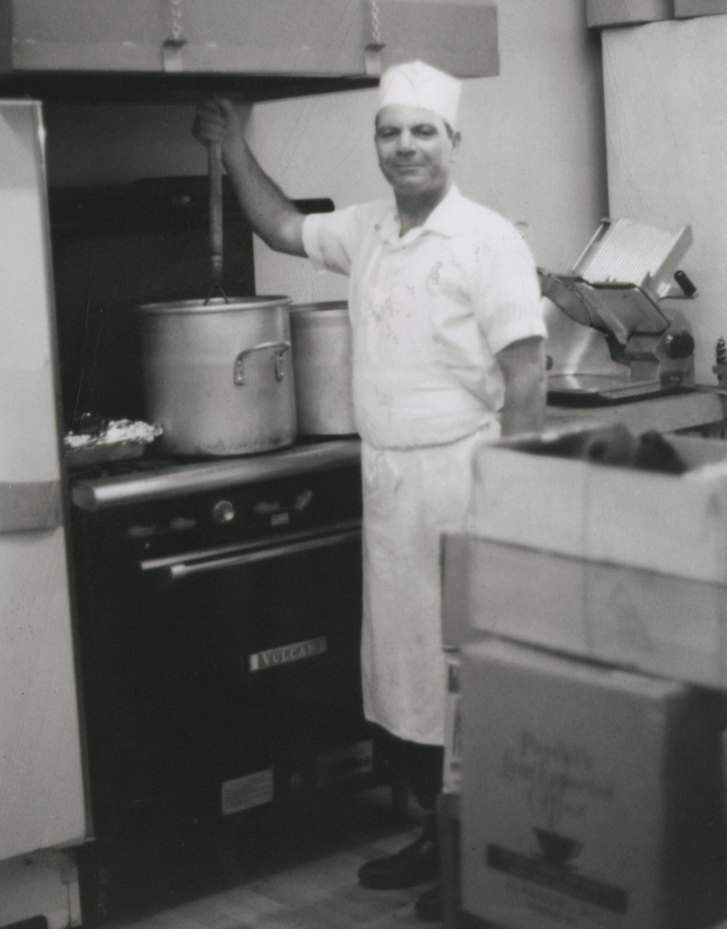 Black and white photo of a male chef in a kitchen, wearing a white apron and chef's hat, stirring a large pot on a stove.