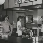 Two women standing behind a counter in a cafe or restaurant, preparing food or drinks.