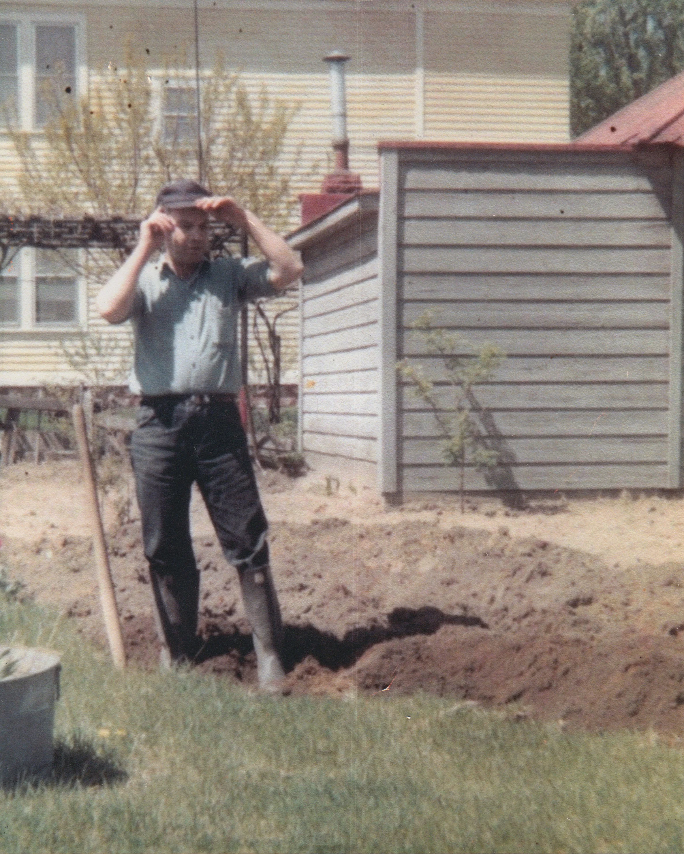 A young man in a gray shirt and dark pants standing in a garden with a shovel in a freshly dug hole, with a house and shed in the background.