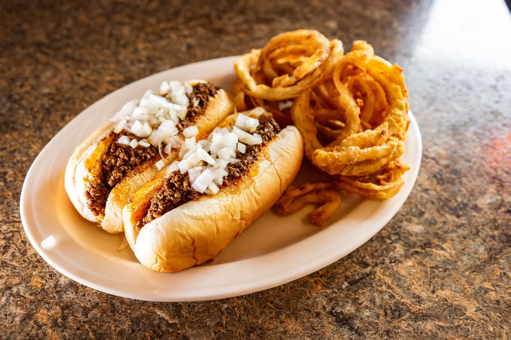 Hot dog with chopped onions and chili, served with onion rings on a white plate.