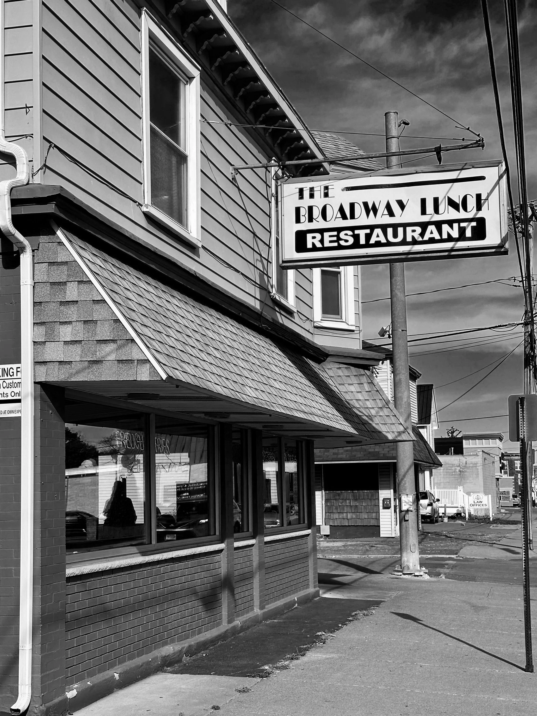 Black and white photo of a restaurant building with a sign that reads "The Broadway Lunch Restaurant" hanging above the sidewalk.