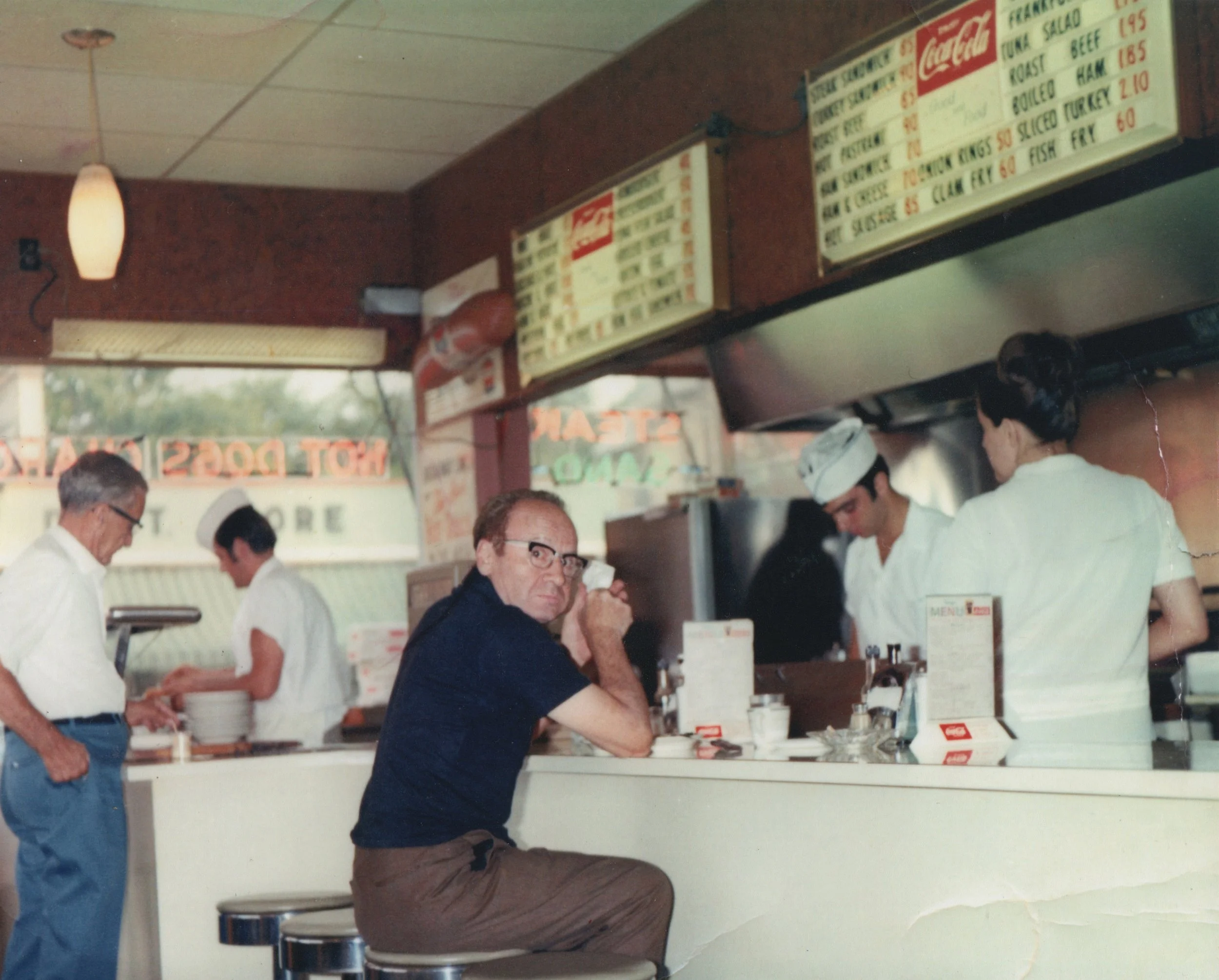A man with glasses sitting at a counter in a retro diner, with two women in white uniforms working behind the counter, and two men, one elderly and one middle-aged, standing nearby. The diner has a menu board and neon signs in the background.