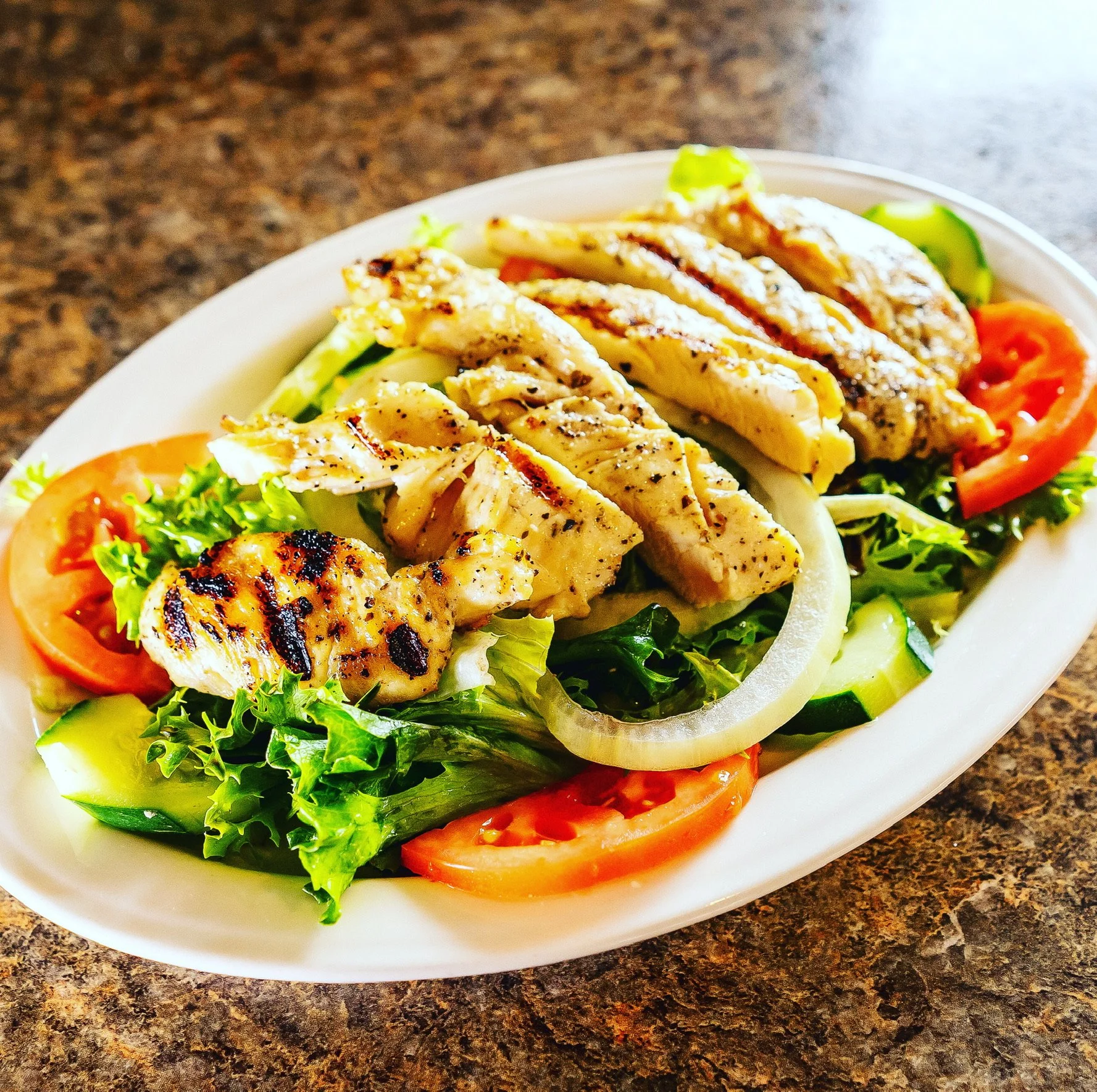 A salad with grilled chicken strips, sliced tomatoes, cucumbers, onions, and leafy greens in a white bowl on a brown countertop.