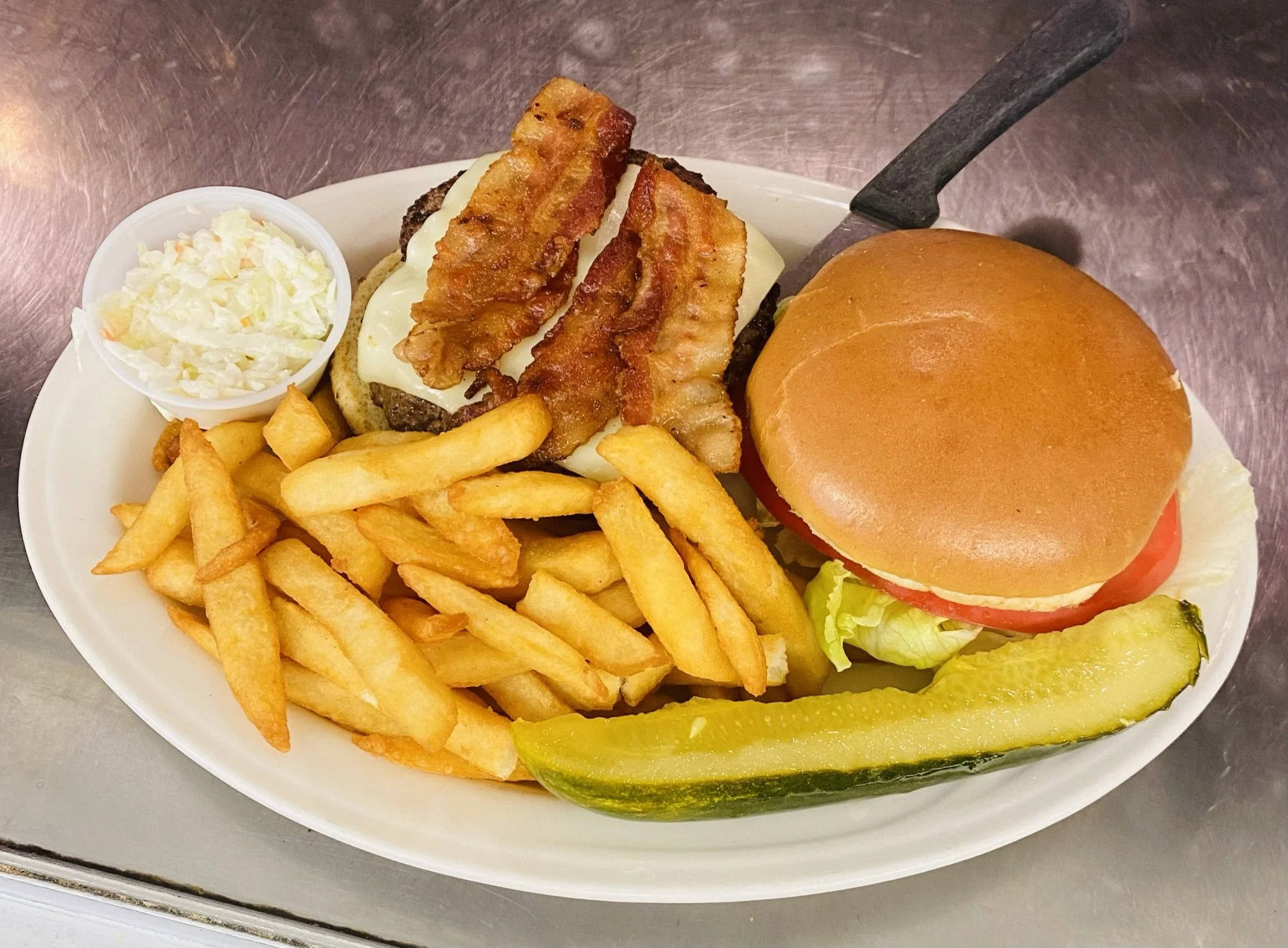 A plate with a cheeseburger topped with bacon, French fries, a pickle spear, a slice of tomato, lettuce, and mayonnaise, and a small cup of coleslaw.