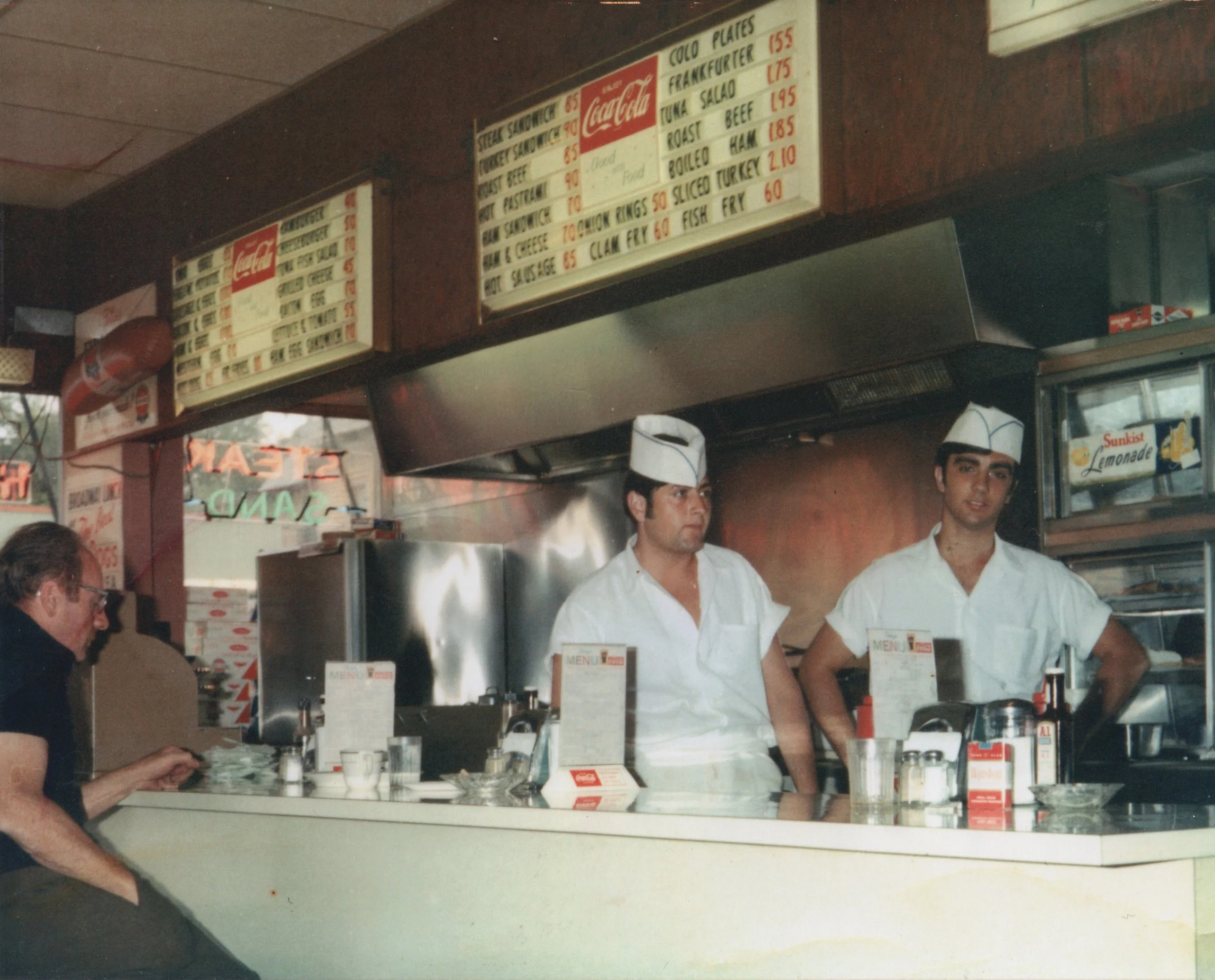 Two men working at a sandwich shop counter wearing white uniforms and hats, with a customer sitting nearby, and a menu board above them listing sandwiches and prices.