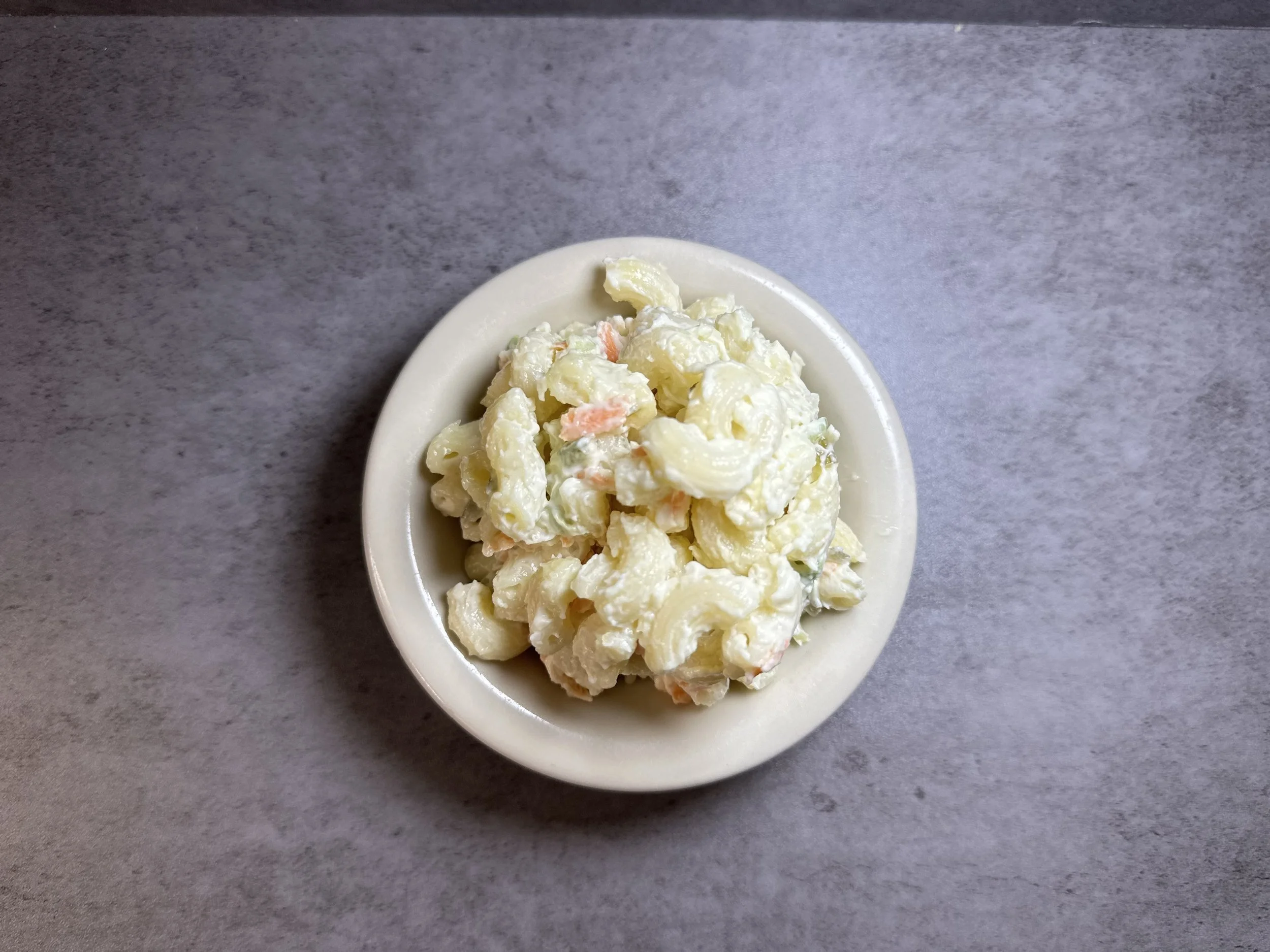 A small white bowl of macaroni salad on a gray surface.