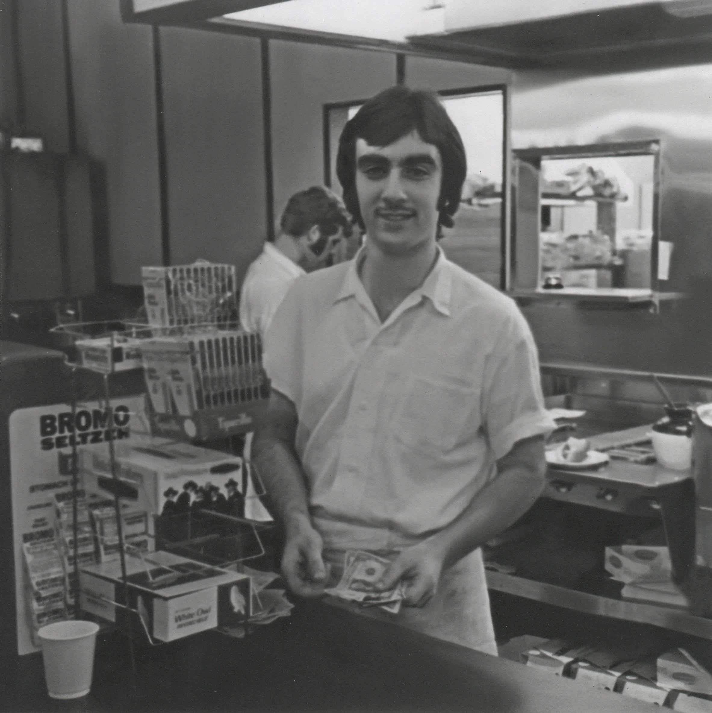 A young man with dark hair and a mustache, wearing a light-colored short-sleeve shirt, is standing behind a counter in a convenience store, counting money. There is another person in the background, and various products and shelves are visible.