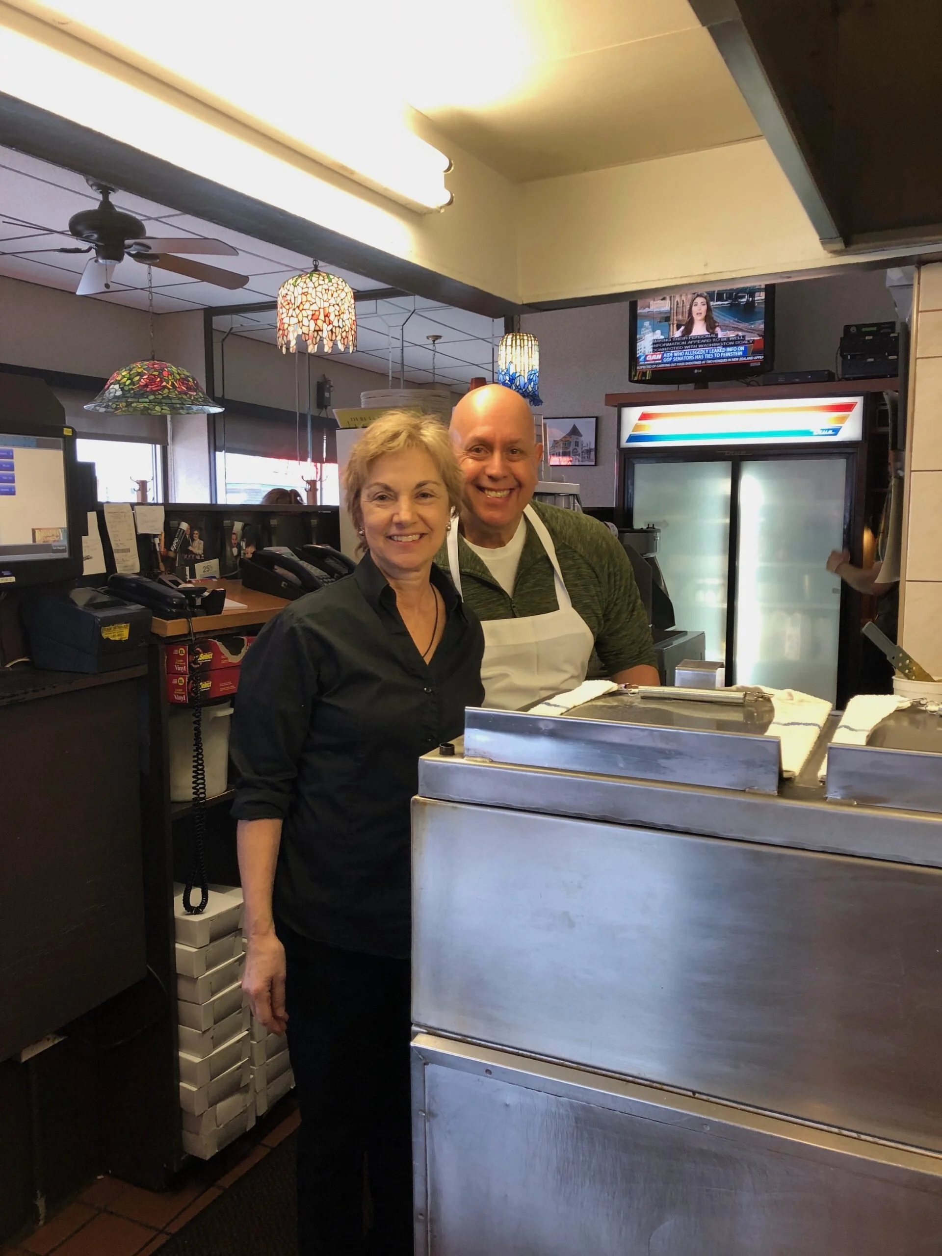 A woman and a man standing behind a counter in a restaurant or diner, smiling at the camera. The woman is wearing a black shirt, and the man is wearing a green shirt with a white apron. The background shows decorative hanging lamps, a TV, and various