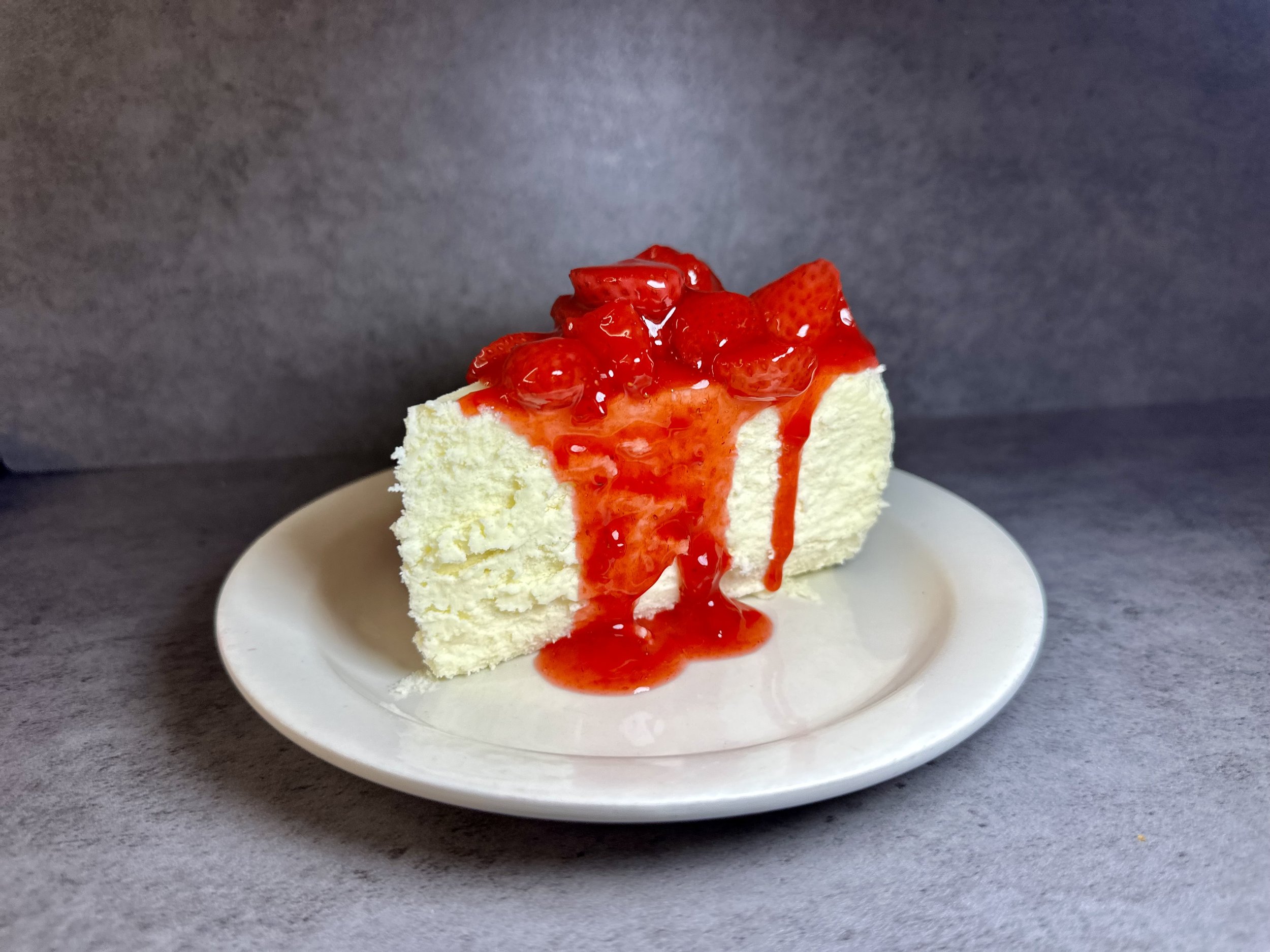 Slice of cheesecake with strawberry topping and strawberry syrup on a white plate against a gray background.
