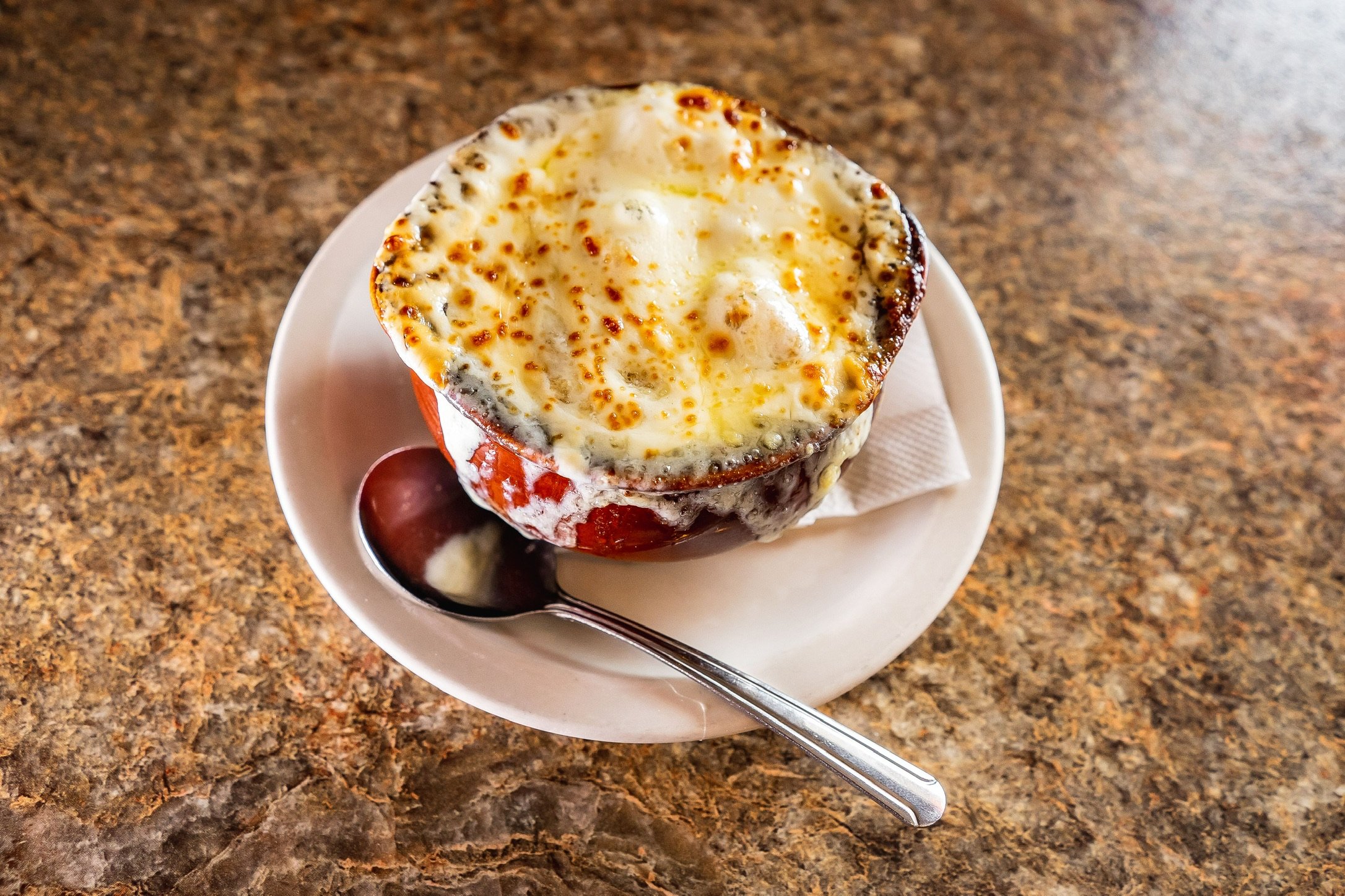 Baked stuffed apple dessert in a bowl with a spoon on a white saucer on a brown speckled countertop.