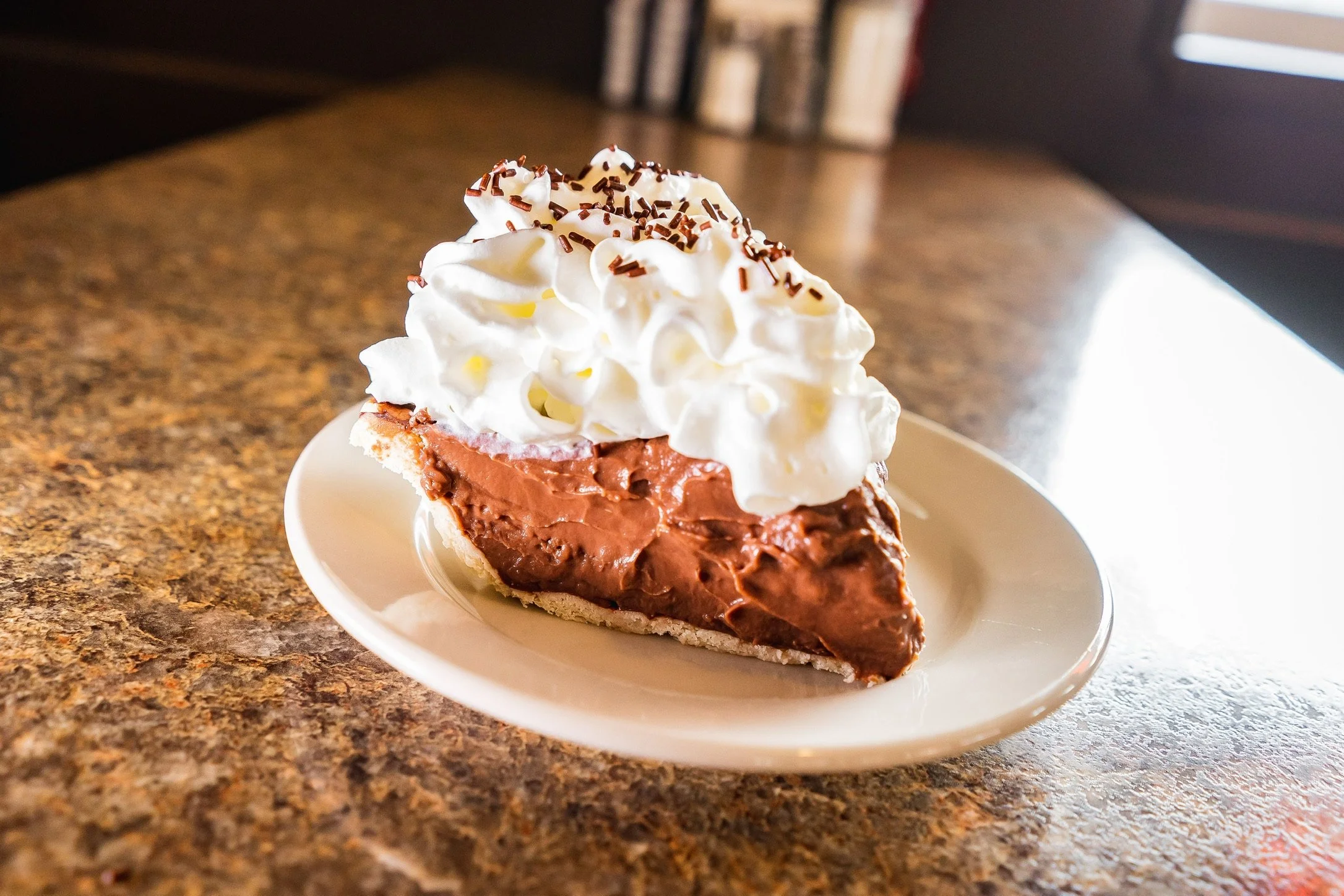 Slice of chocolate cream pie with whipped cream and chocolate sprinkles on a white plate.