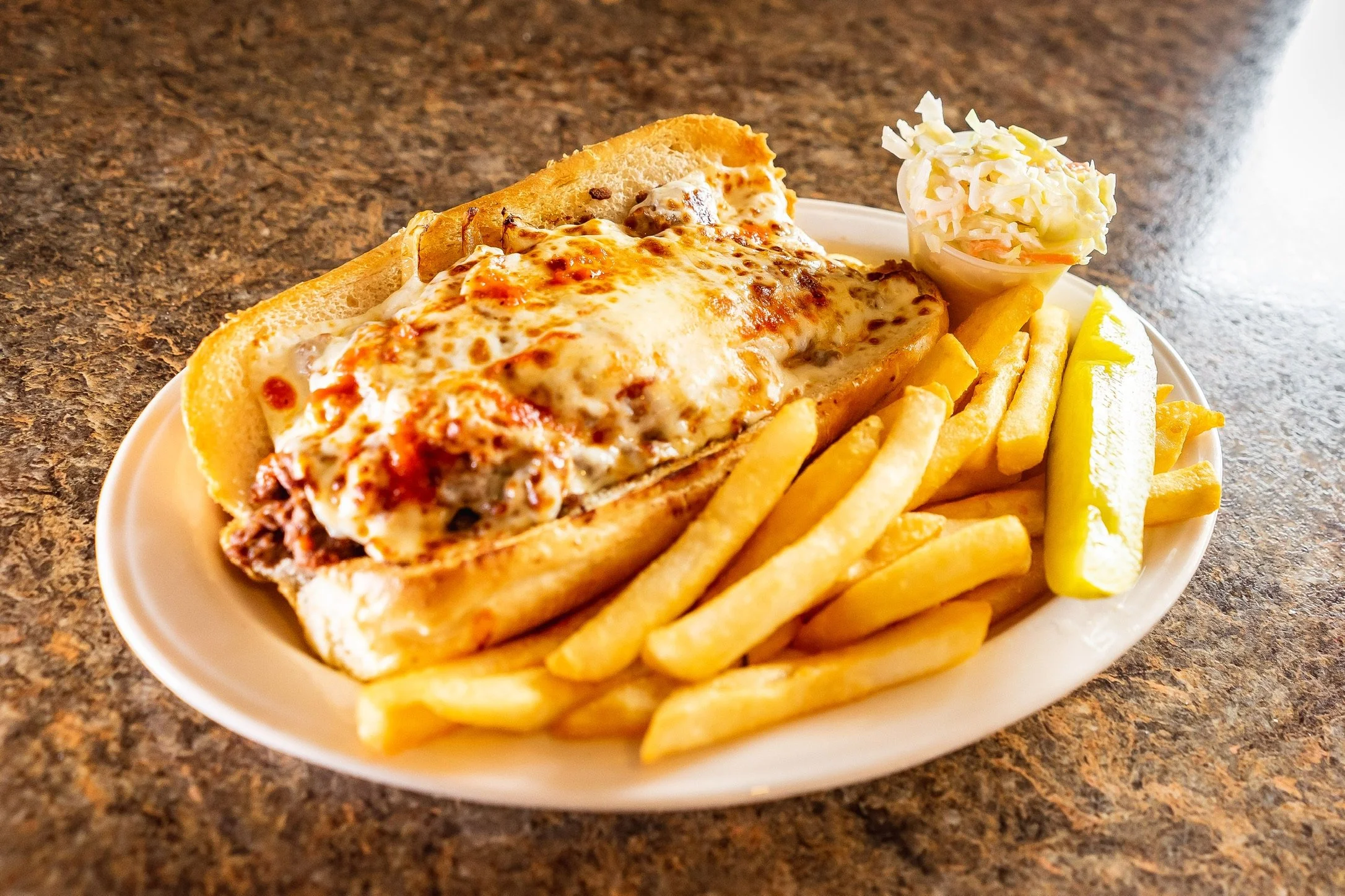 Cheeseburger sub with melted cheese, served with French fries, pickles, and coleslaw on a white plate.