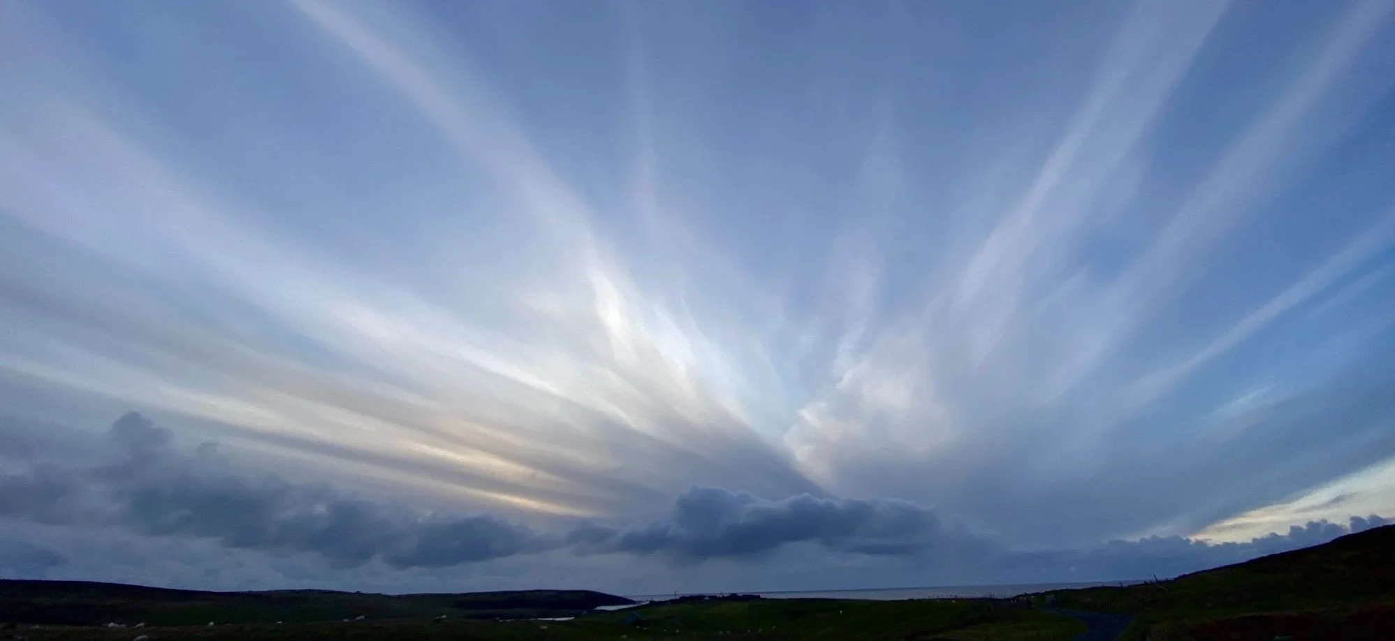 A vast open landscape with green grass and a body of water in the distance under a dramatic sky filled with elongated, wispy clouds.
