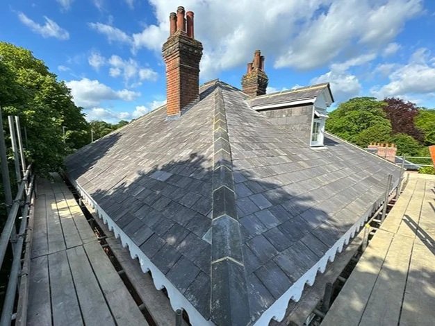 Rooftop of a house with slate tiles, two brick chimneys, and a small dormer window under a blue sky.