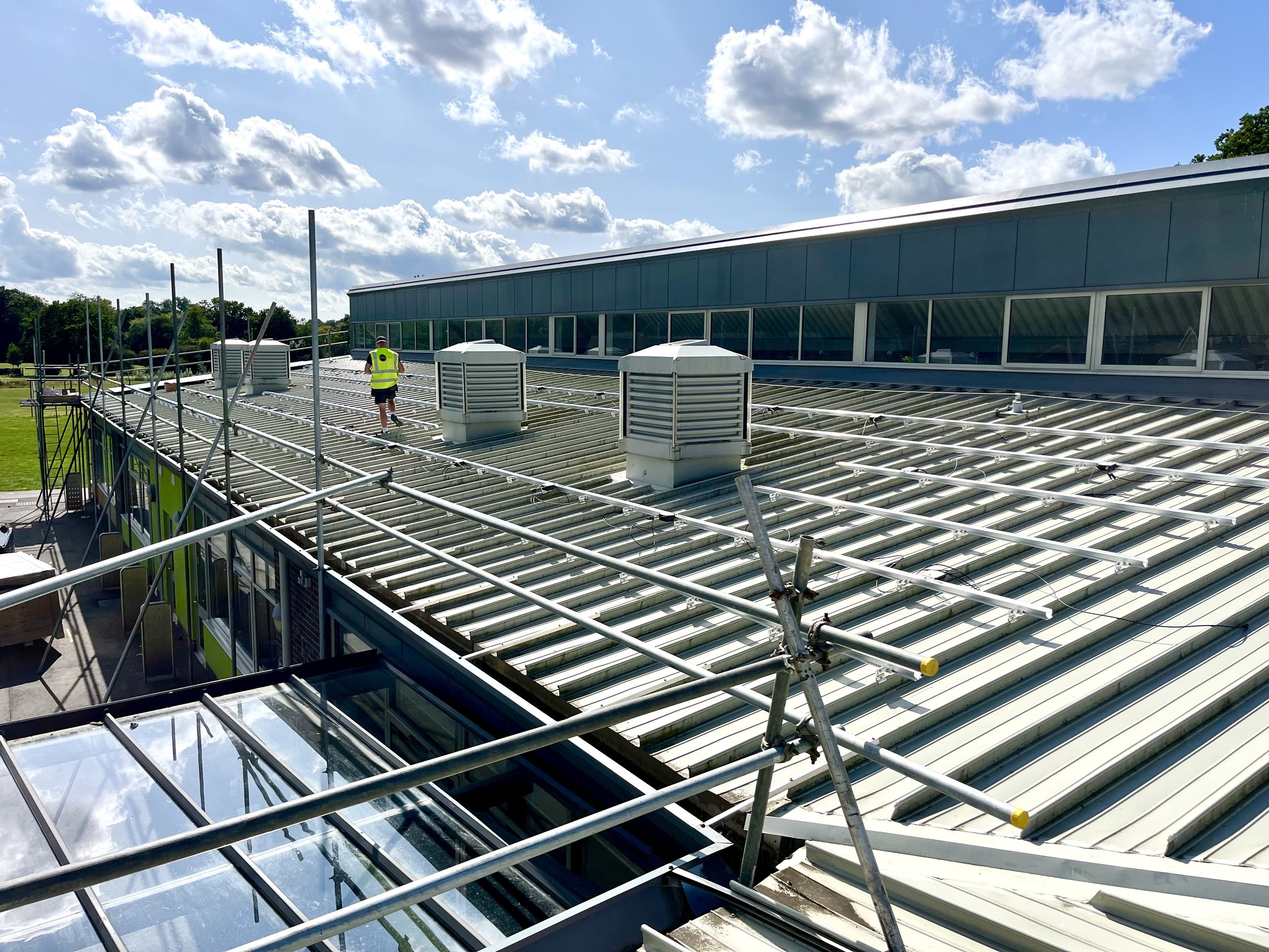 A person inspecting a rooftop with metal rails, vents, and scaffolding on a sunny day with scattered clouds.