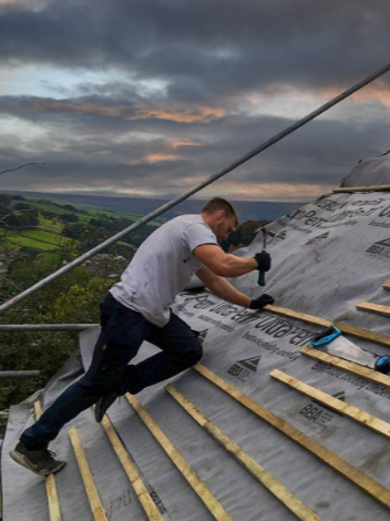 A man working on a roof, installing wooden battens over a weatherproofing membrane, with a scenic landscape and cloudy sky in the background.