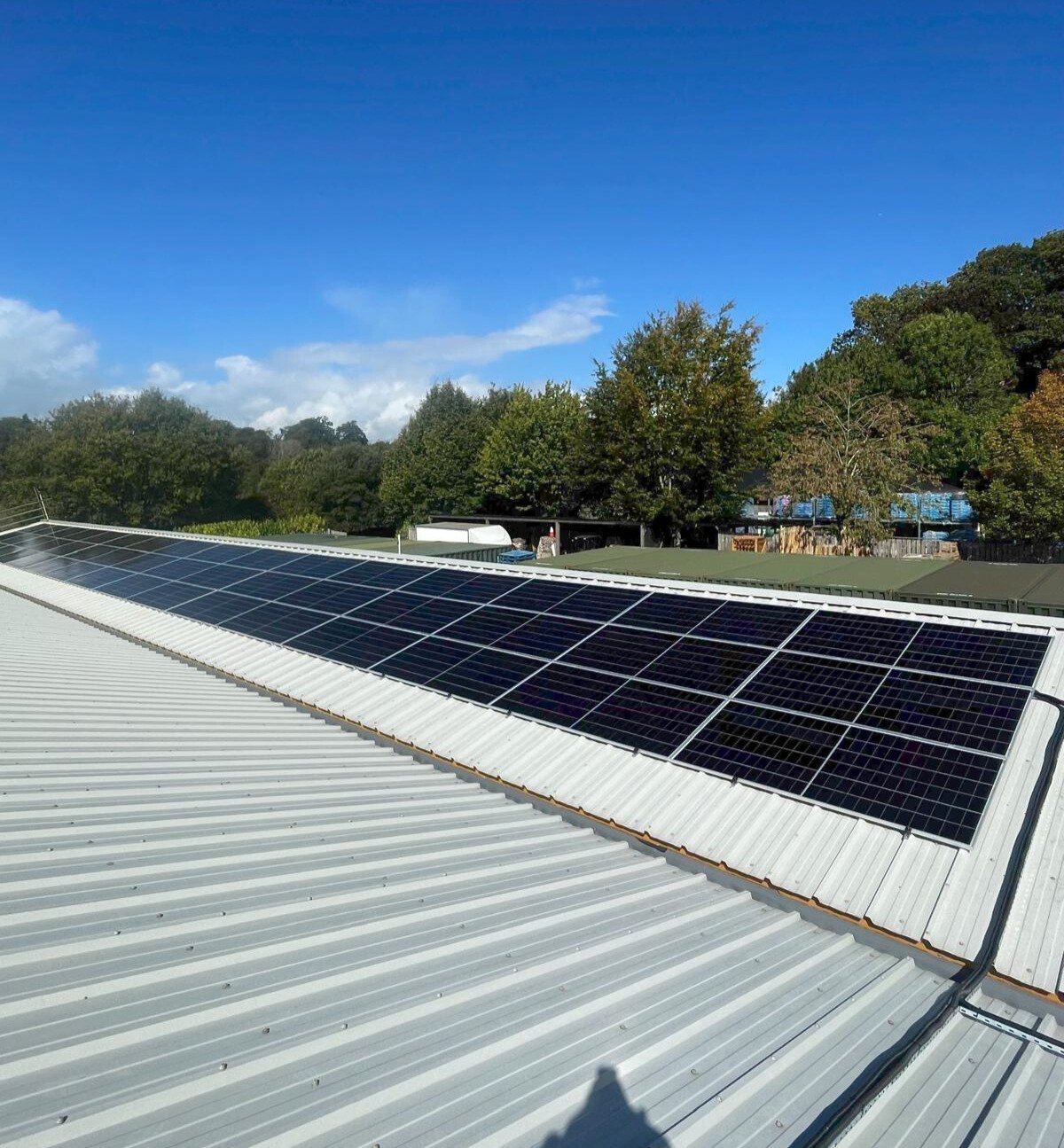 Solar panels installed on a metal rooftop with trees and a bright blue sky in the background.