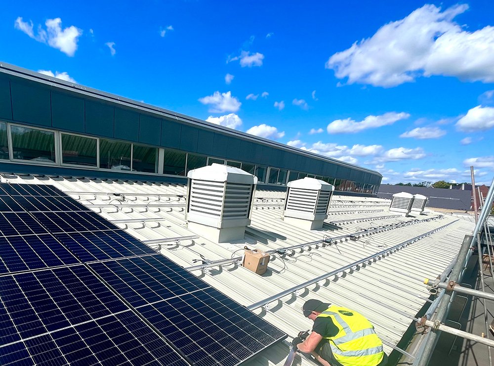 A worker in a yellow safety vest and black shirt installing solar panels on a rooftop with a blue sky and scattered clouds in the background.