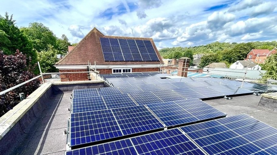 Multiple solar panels installed on a rooftop of a residential building with a brick house and green trees in the background.