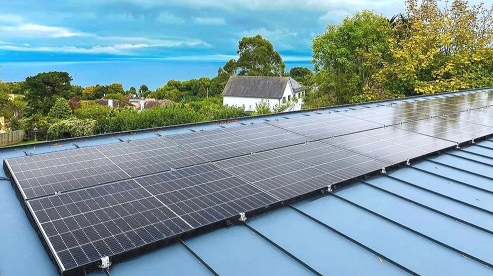 Solar panels installed on a blue metal roof with a scenic view of green trees and houses under a partly cloudy sky.