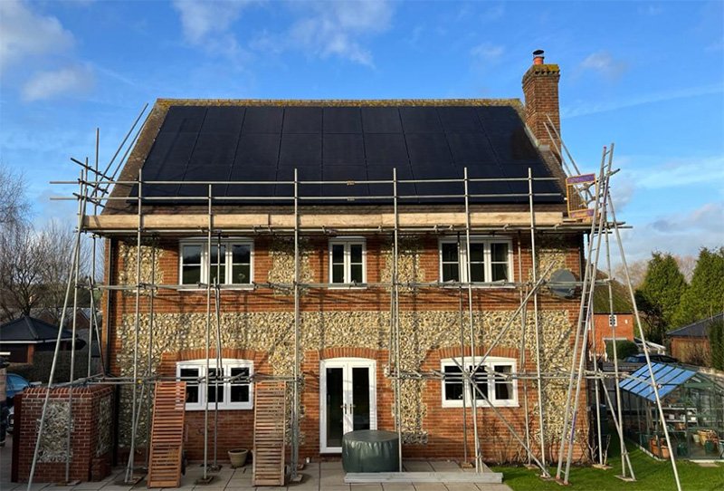 A brick and stone house under renovation with scaffolding surrounding it. The house has a black roof, several windows, and a chimney. Construction materials are visible in the front yard.