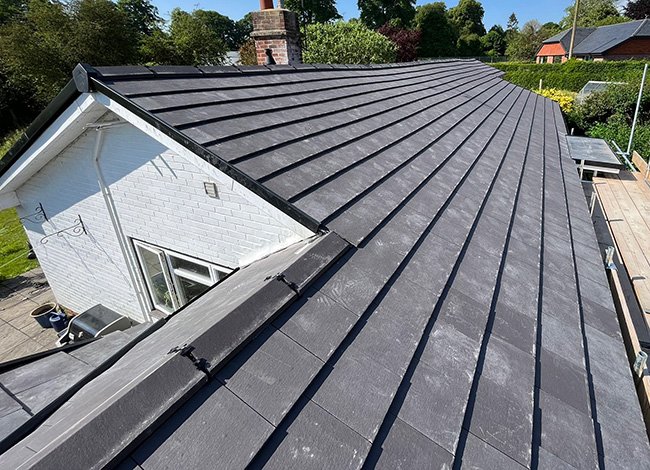 View of a black metal roof on a residential house, showing shingles and a chimney in the background, surrounding trees, and a partially visible deck.