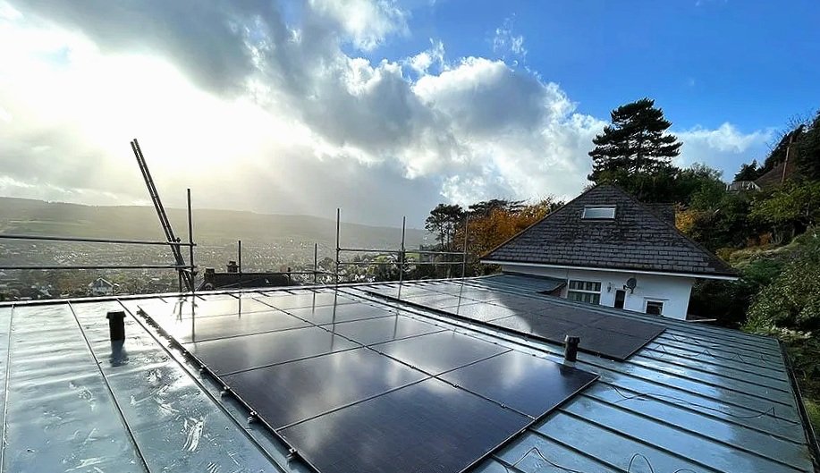 View of a house's roof with solar panels, surrounded by trees and a cloudy sky in the background.