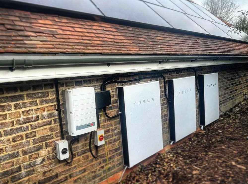 Exterior view of a brick house wall with four Tesla power wall batteries and solar panels on the roof.
